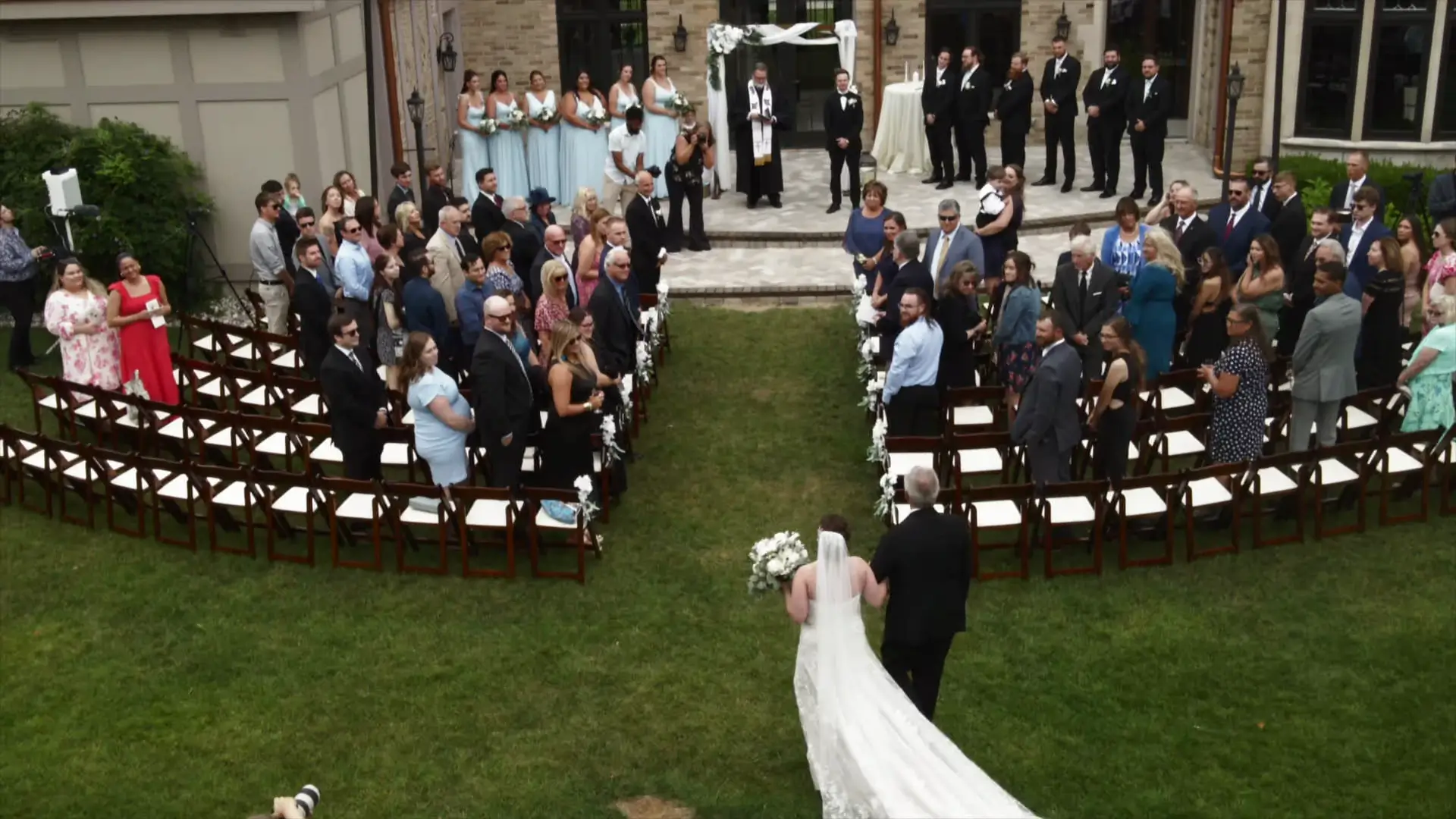 Aerial view of Leah walking down the aisle at Venue3Two in Grand Rapids, Michigan, captured by drone. Guests are standing and the wedding party is assembled at the altar. The outdoor ceremony creates an elegant, sentimental atmosphere.