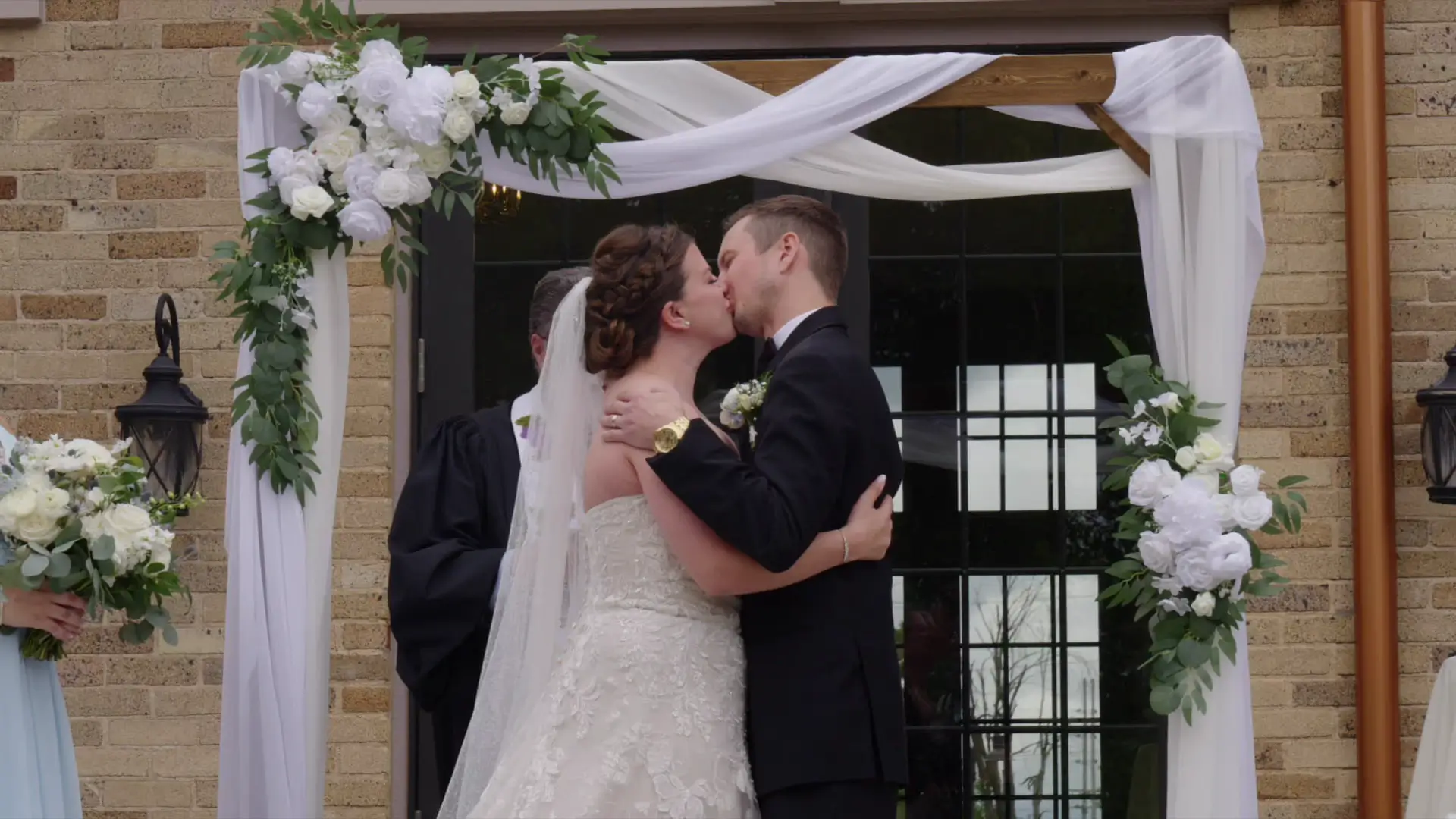 In this wedding highlight video still, Leah and Eric share their first kiss under a floral arch at Venue3Two in Grand Rapids, Michigan. The videographer captures this romantic moment as they embrace, surrounded by elegant white flowers and drapery.