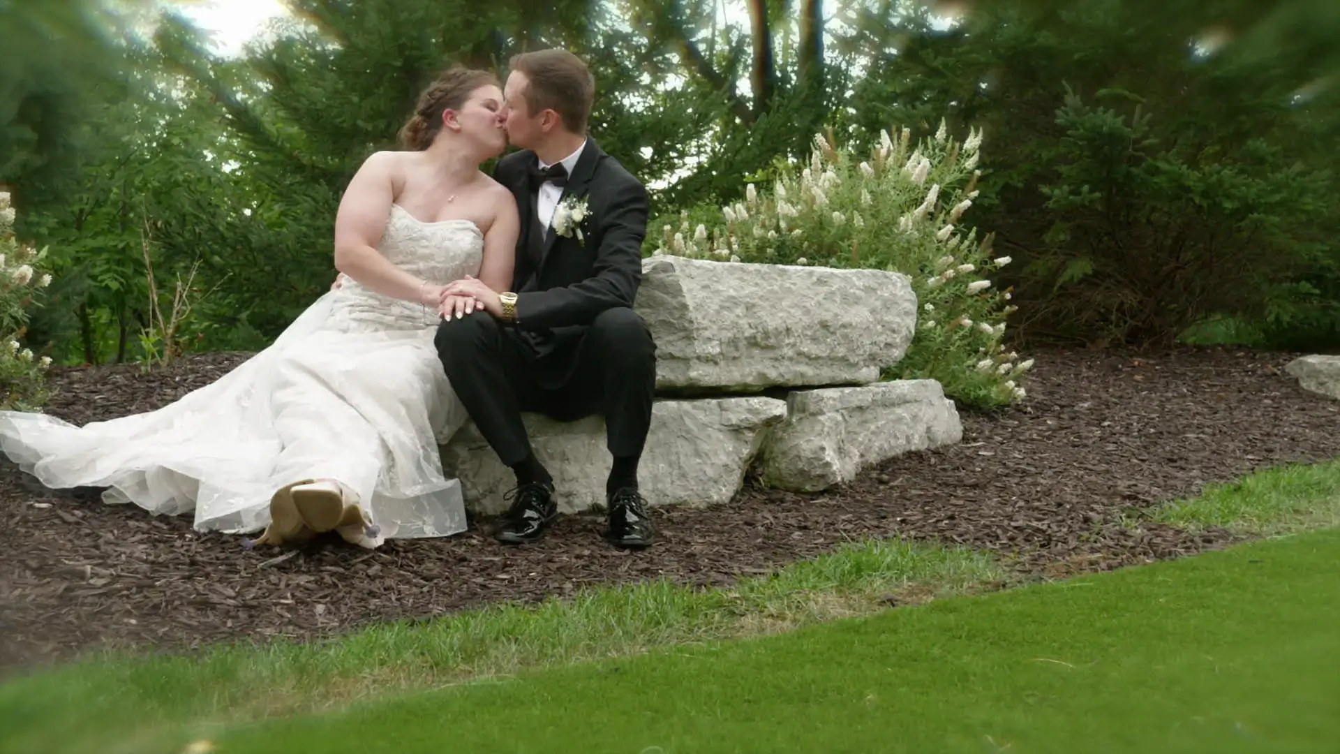 In this snapshot from their wedding highlight, Leah and Eric share a kiss on a stone bench surrounded by lush Michigan greenery at Venue3Two, captured beautifully by their wedding videographer.