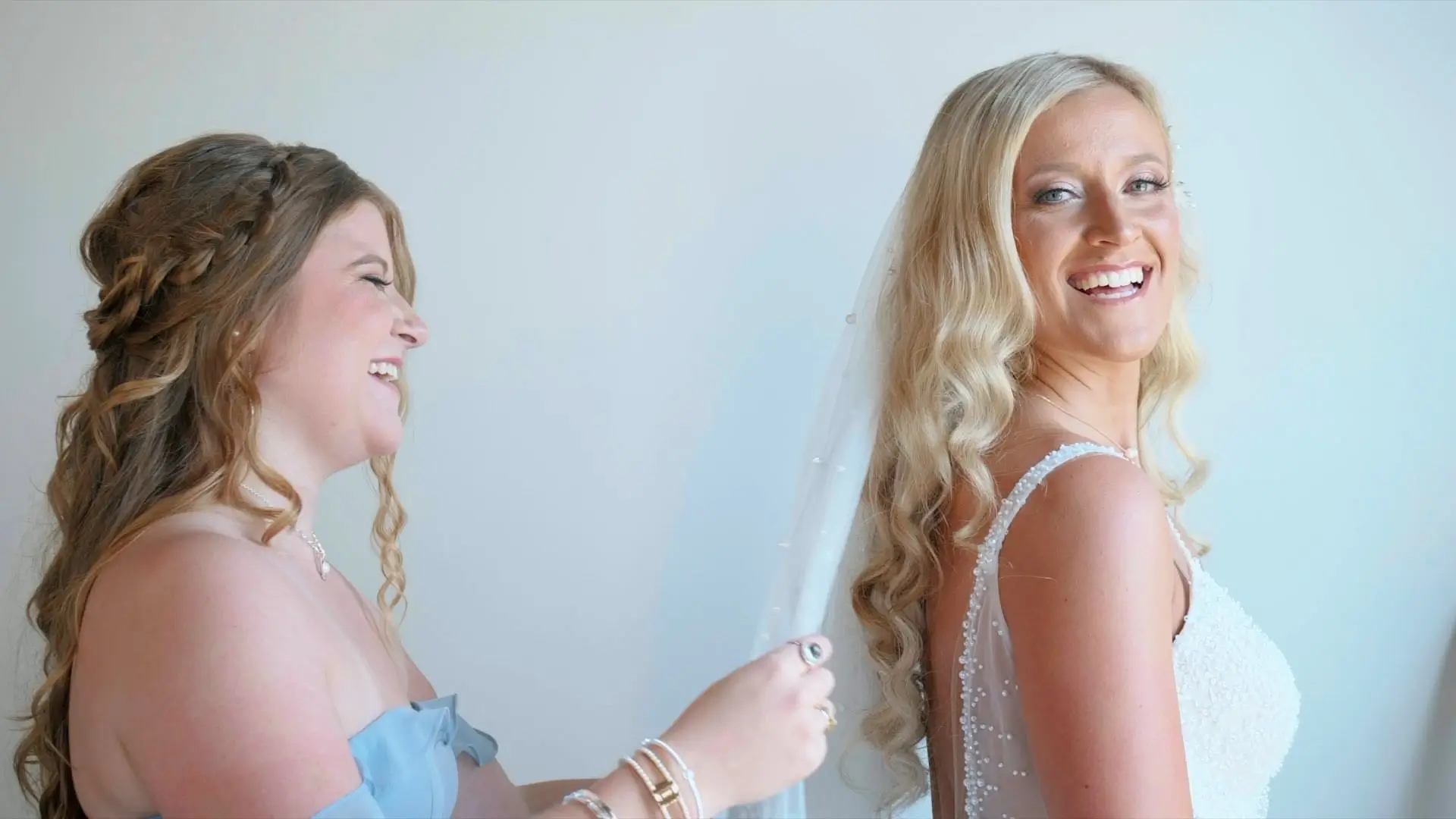 A smiling bride, Kasey, in a white wedding dress, is having her veil adjusted by a bridesmaid in a light blue dress. Both women have long, wavy hair and are laughing joyfully in a bright room.