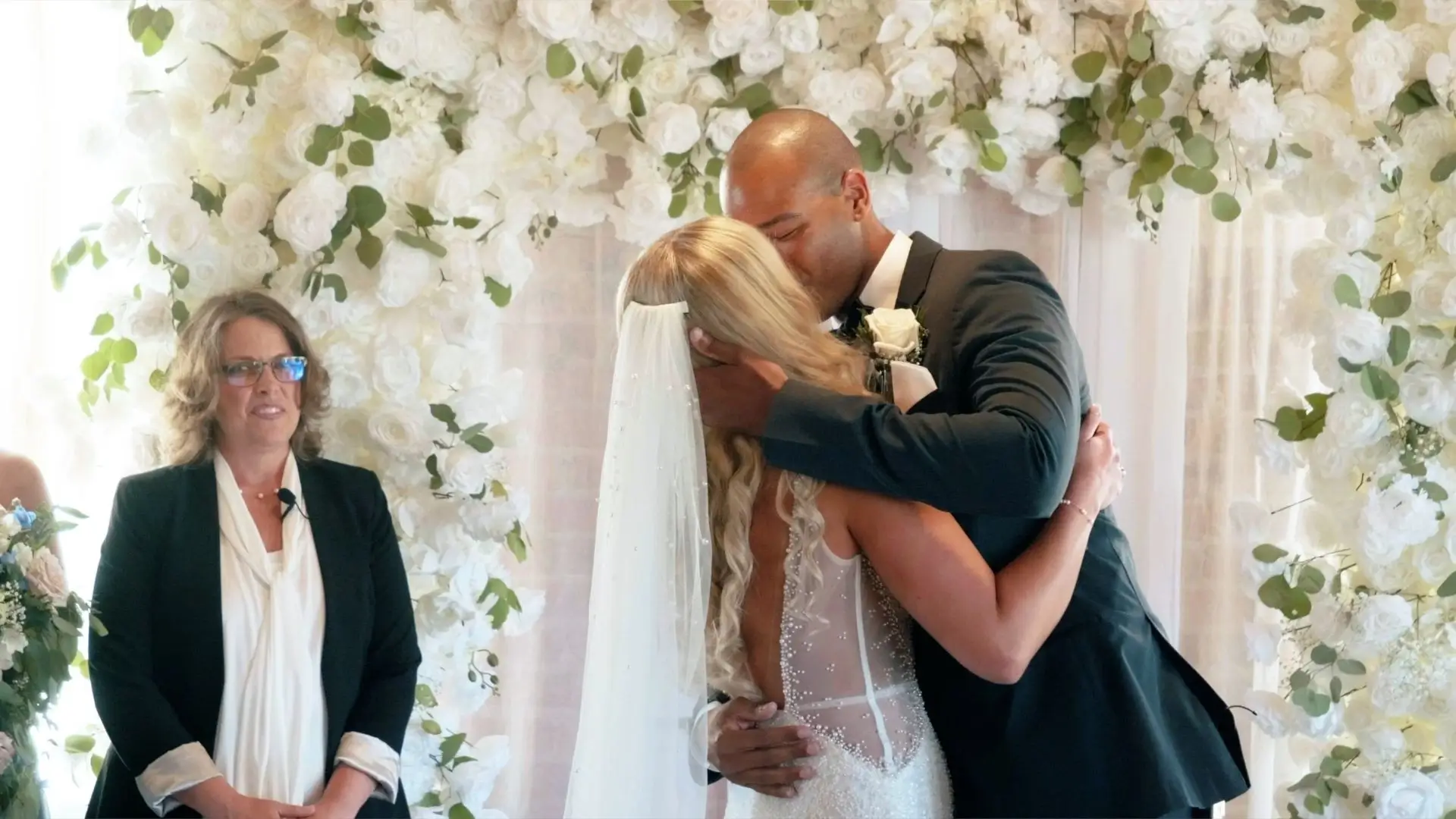Roberto and Kasey share their first kiss as a married couple at the Record Box Loft in Battle Creek, surrounded by a beautiful white floral arch. The officiant stands nearby, smiling warmly at the newlyweds.