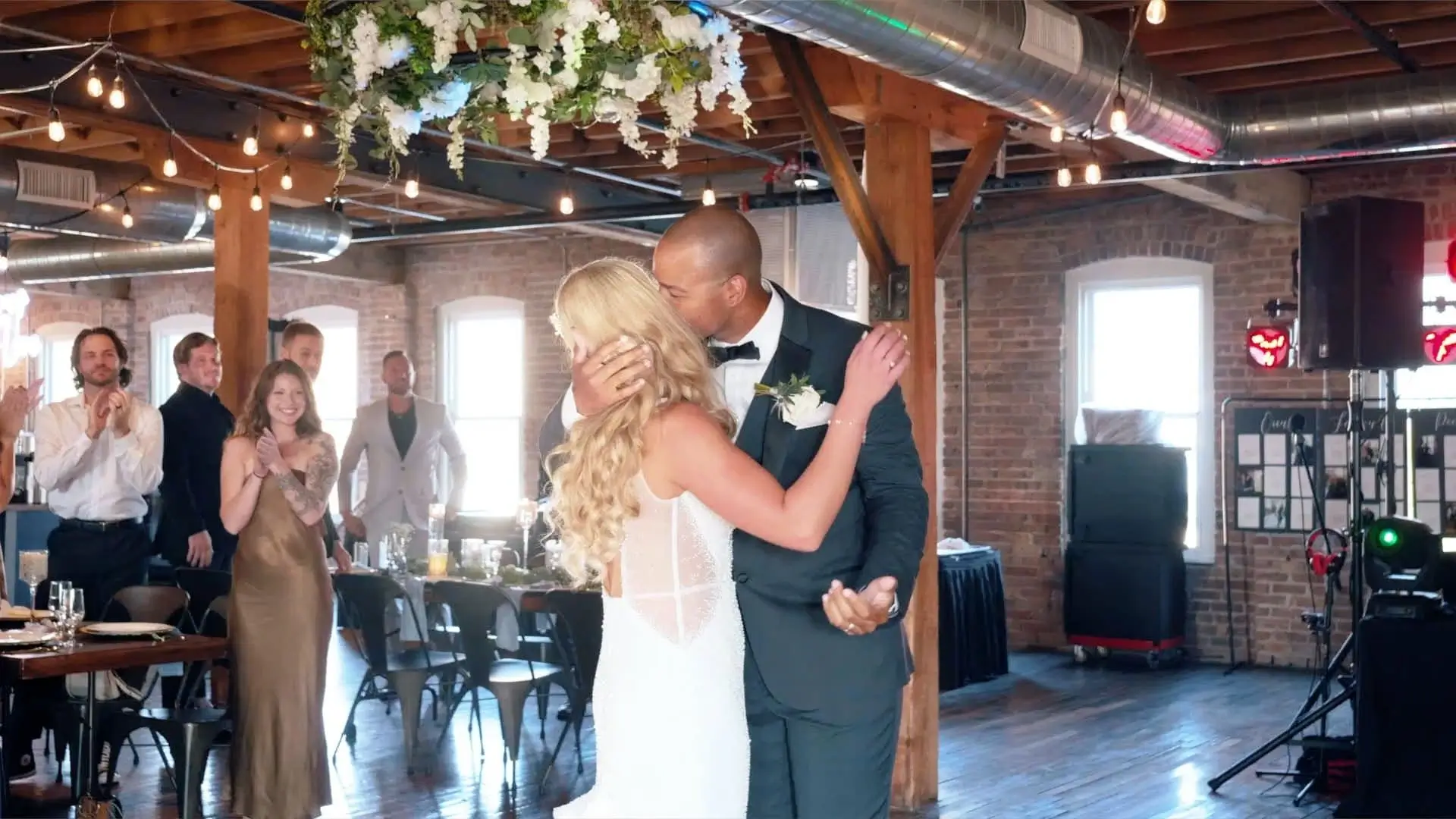 In this still from their wedding video at the Record Box Loft in Battle Creek, Roberto and Kasey share a loving kiss during their reception as guests clap and smile in the background, beautifully capturing the newlyweds' joyful celebration.