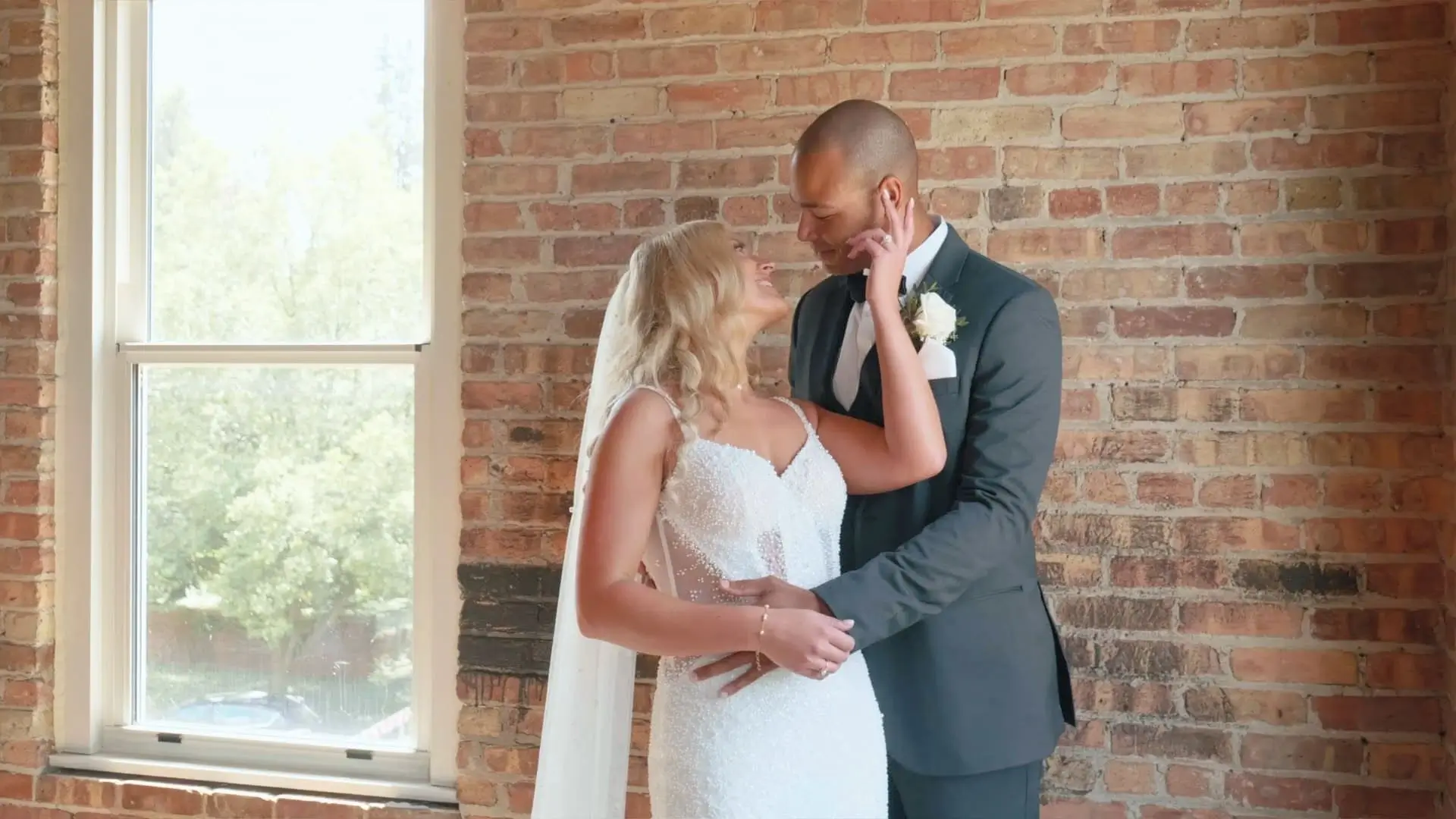 Bride Kasey and groom Roberto stand close and share a loving gaze in front of a brick wall at the Record Box Loft in Battle Creek, Michigan. This moment from their wedding video captures their playful and affectionate demeanor on July 13, 2024.