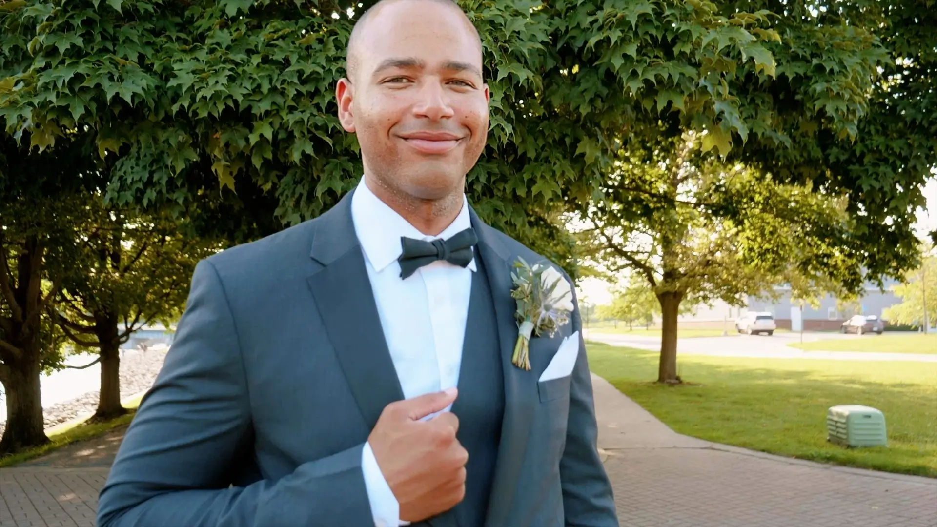 In this still from their wedding video, Roberto is seen smiling confidently while adjusting his suit, standing in front of lush greenery. The setting is the Record Box Loft in Battle Creek, Michigan