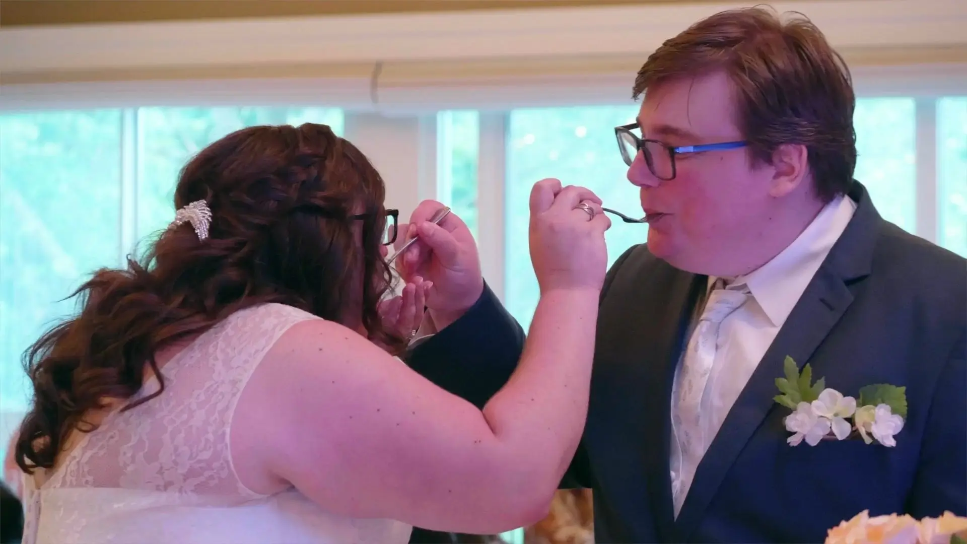 Nichole and Seth feeding each other with spoons at their wedding reception at Ann Arbor City Club. Nichole is in a lace wedding dress with a hair accessory, and Seth is in a suit with a floral boutonniere.