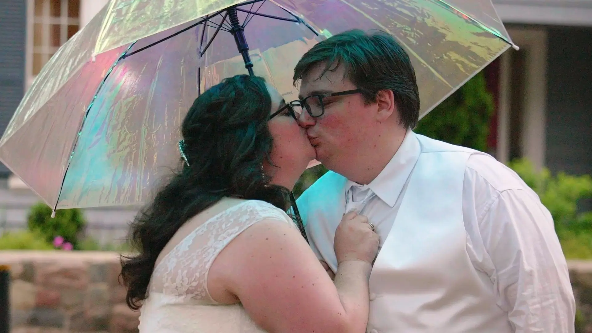 Nichole and Seth share a kiss under a colorful, reflective umbrella. They are dressed in wedding attire; Nichole is in a white lace dress, and Seth is in a white shirt with a tie and vest. They are standing outside the Ann Arbor City Club.