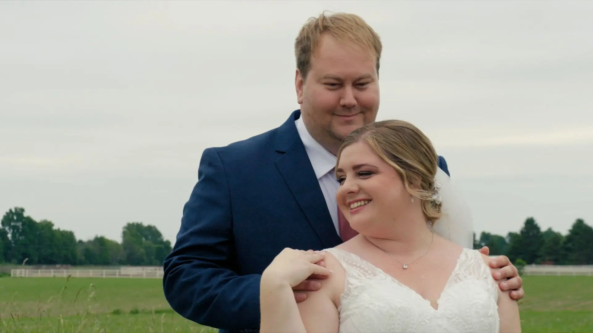  Hannah and Alex smiling on their wedding day at Cherry Blossom Barn. Hannah is wearing a white lace wedding dress and veil, while Alex is in a dark blue suit. They are standing outside in front of a green field with trees and fences in the background.