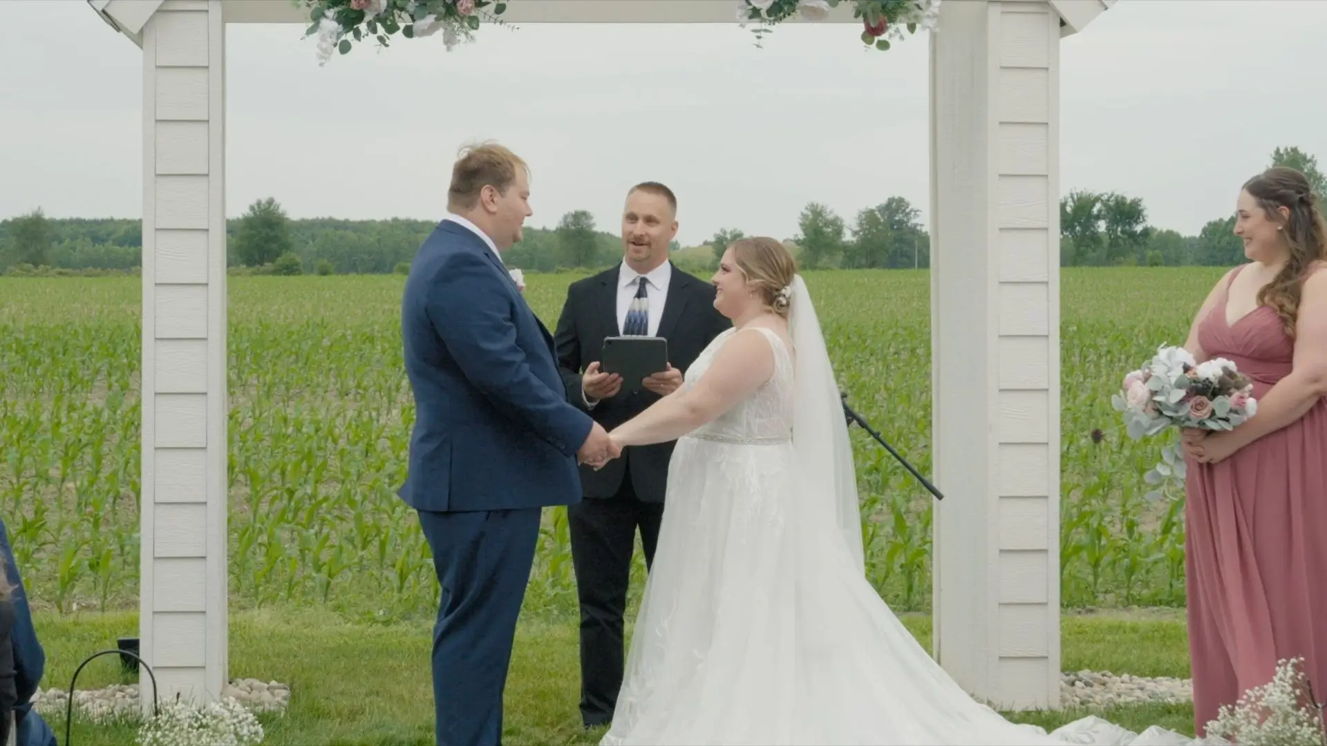Hannah and Alex holding hands and smiling during their outdoor wedding ceremony at Cherry Blossom Barn, with the officiant standing behind them and a bridesmaid holding a bouquet on the side. The ceremony is set against a backdrop of greenery and farmland.