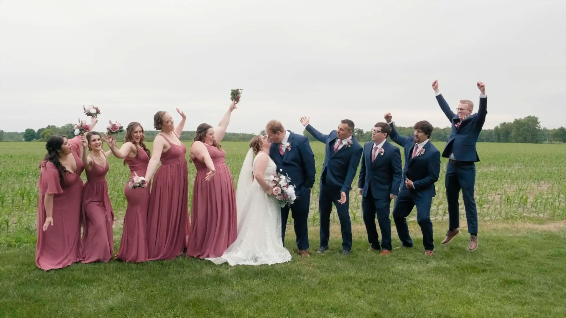 A wedding party celebrates outside in a field at Cherry Blossom Barn. The bride, holding a bouquet, kisses the groom in the center of the group. Bridesmaids in matching dusty rose dresses and groomsmen in navy suits with pink ties cheer around the couple.