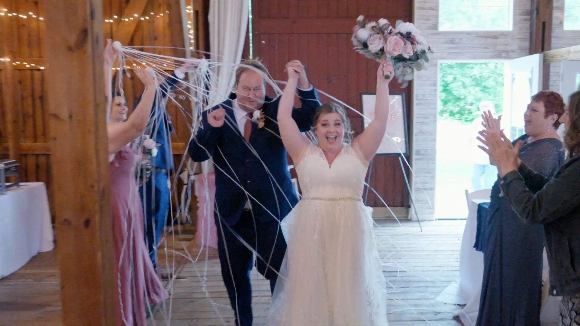 Bride and groom, Hannah and Alex, joyfully enter their reception at a rustic & charming barn, surrounded by guests and streamers. The bride holds a bouquet and both have big smiles, celebrating their union.
