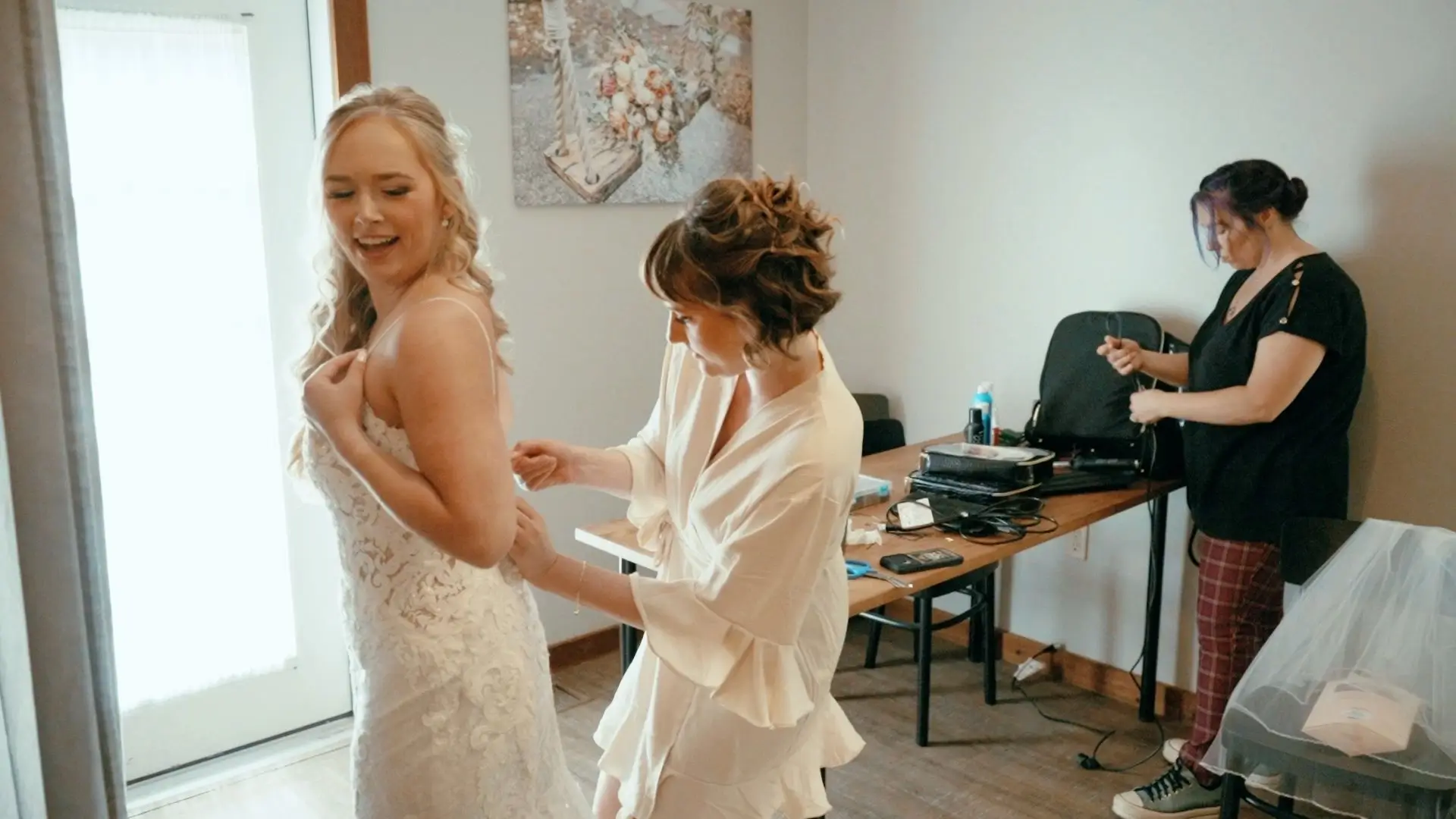 
Bride Caroline smiles joyfully as she gets ready for her wedding at the BlueBridge Events Centre in Grawn. A bridesmaid helps adjust her dress, while a makeup artist prepares nearby.