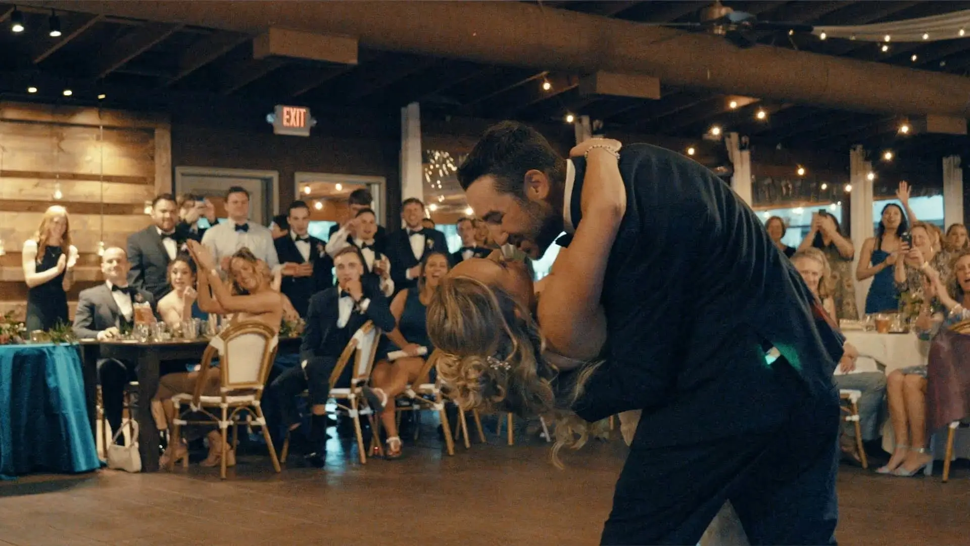 Caroline and Matt share a romantic dip during their first dance at BlueBridge Events Centre in Grawn, Michigan. They are surrounded by cheering friends and family. The atmosphere is festive with guests clapping and capturing the moment on phones.