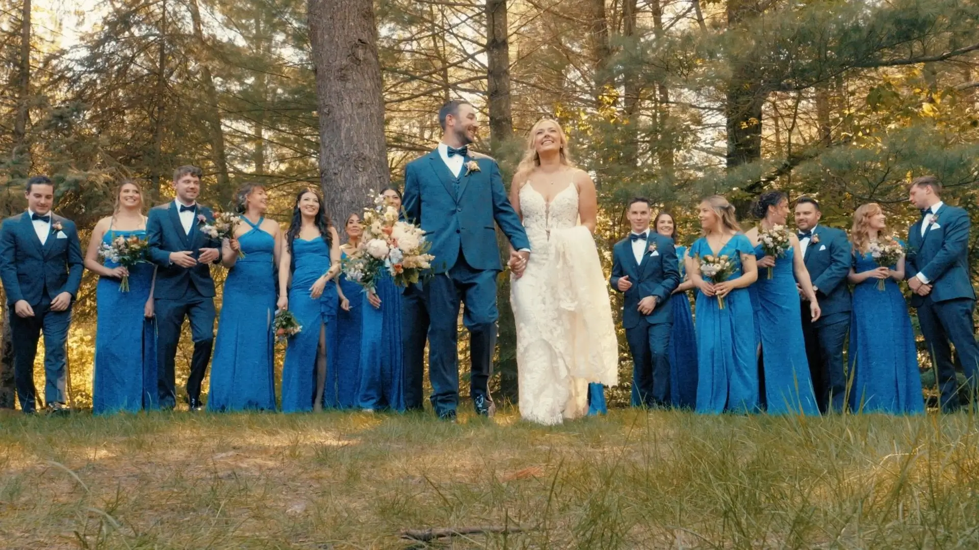 Caroline and Matt stand hand-in-hand, smiling amidst lush Michigan greenery, surrounded by their bridal party in blue attire. The forested backdrop creates a serene and enchanting atmosphere.