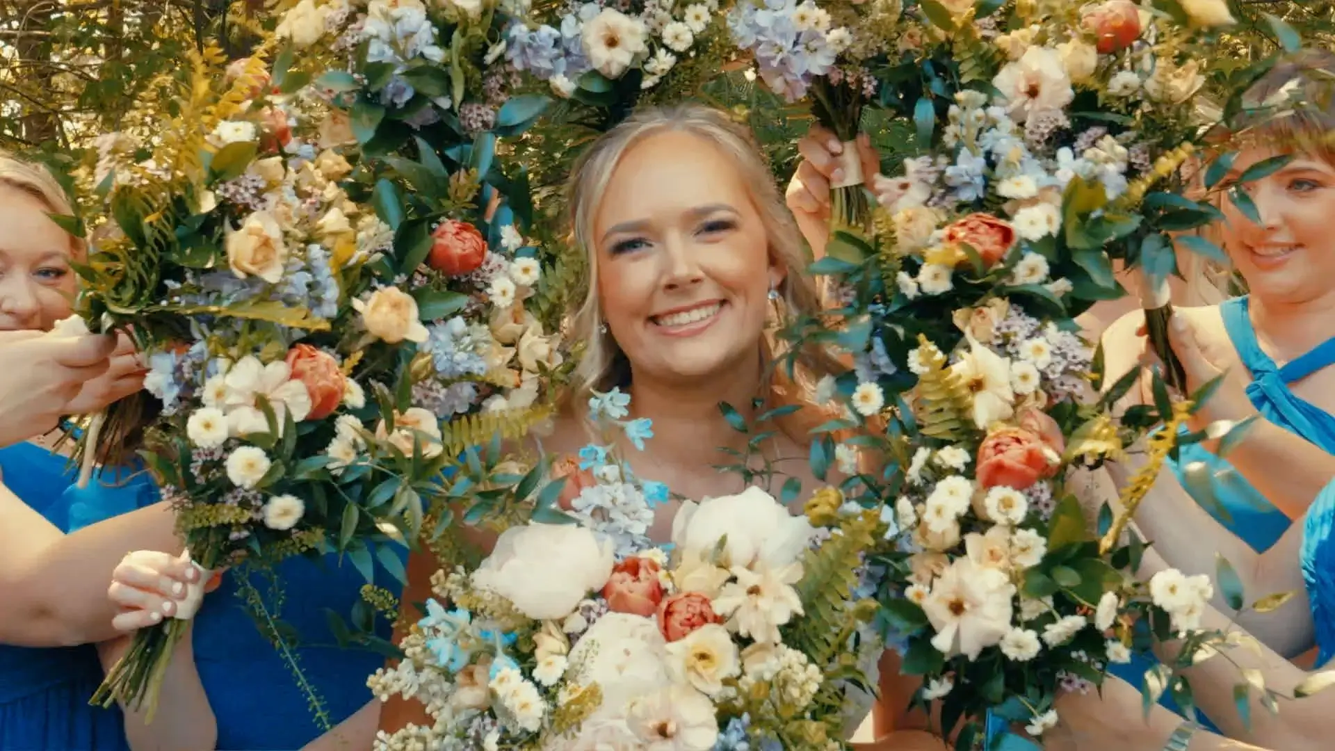 Bride Caroline smiling through a circular arrangement of colorful flowers held by bridesmaids in blue dresses. The vibrant display enhances her joyful expression. The image captures a cheerful moment from the wedding at BlueBridge Events.