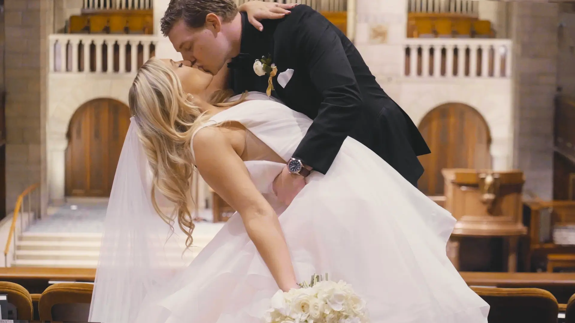 A bride and groom share a romantic kiss inside a church in downtown Grand Rapids, Michigan. The groom, dressed in a black tuxedo, dips the bride, who holds a bouquet of white roses and wears a flowing white gown and veil. 