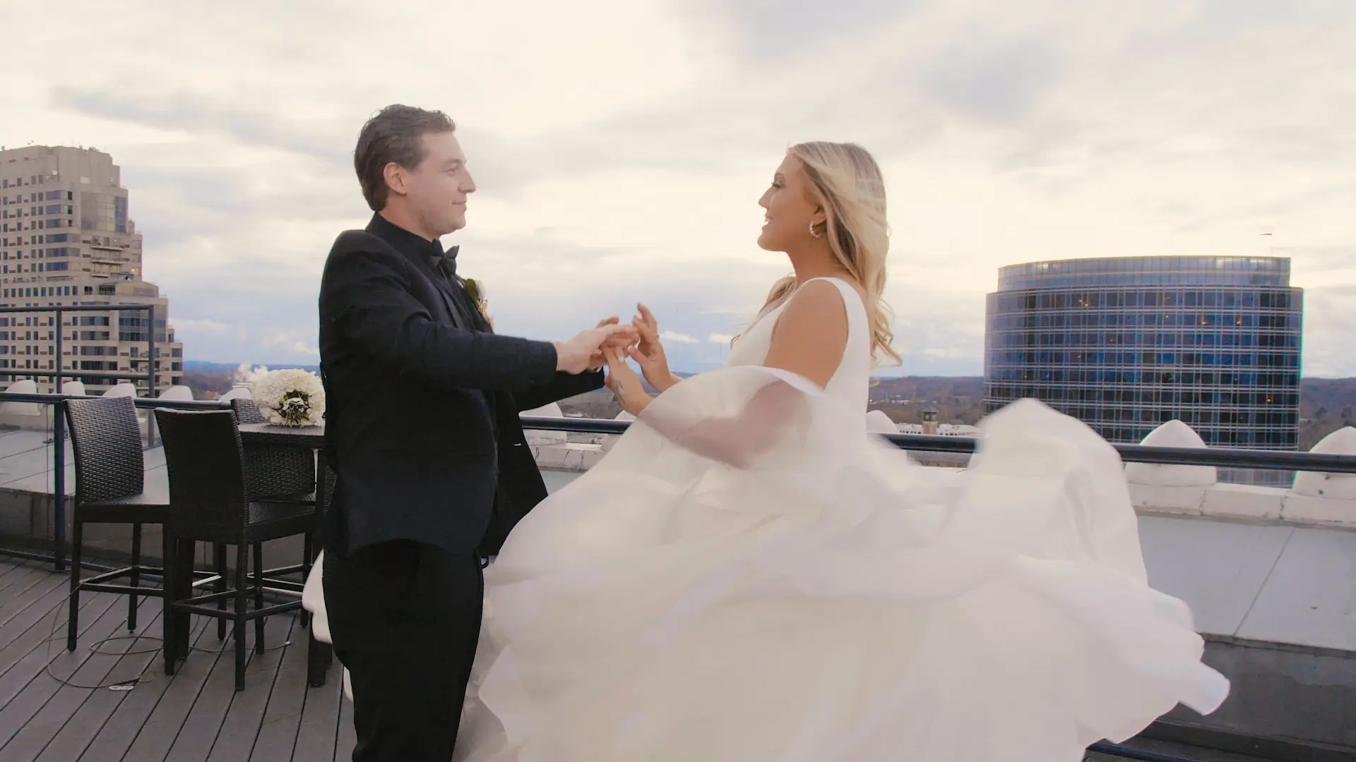 Alyssa and Shane dance on a rooftop in downtown Grand Rapids, Michigan, with city buildings and a cloudy sky in the background. She is in a flowing white wedding dress, and he is in a black tuxedo, matching the wedding's elegant theme.