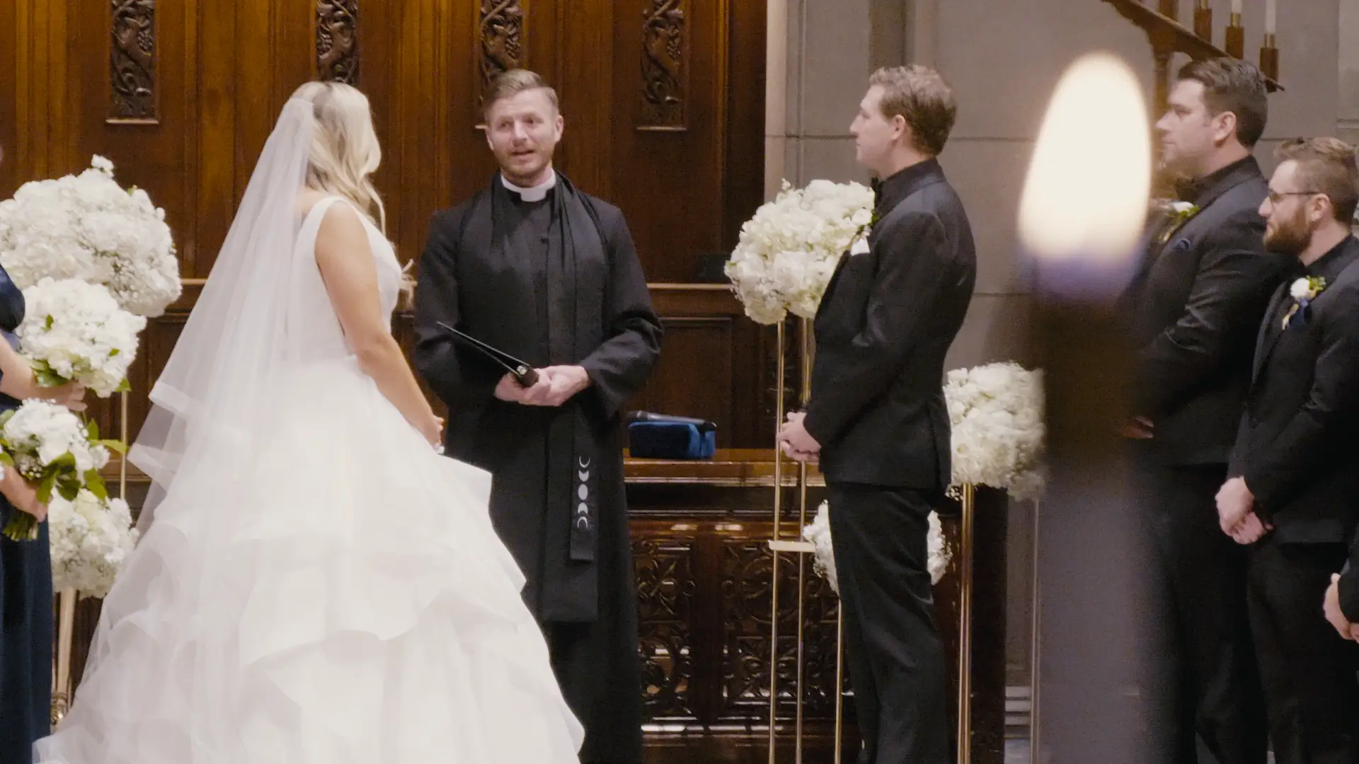 The bride and groom, Alyssa and Shane, stand before an officiant during their wedding ceremony. The bride wears a white dress and holds a bouquet, while the groom and groomsmen are in black suits. Elegant floral decor adorns the venue.