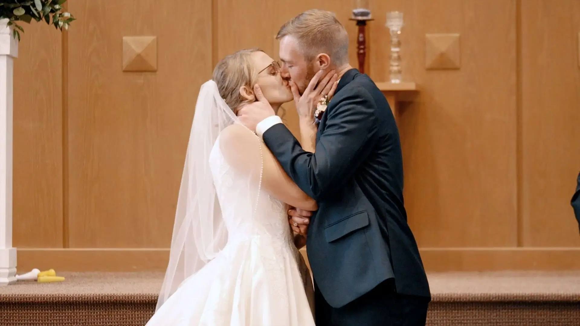 Newlyweds Julia and Andrew share a kiss during their wedding ceremony at 14th Street Church in Holland. Julia, in a white gown and veil, and Andrew, in a dark suit, embrace each other tenderly against the church’s wooden backdrop.