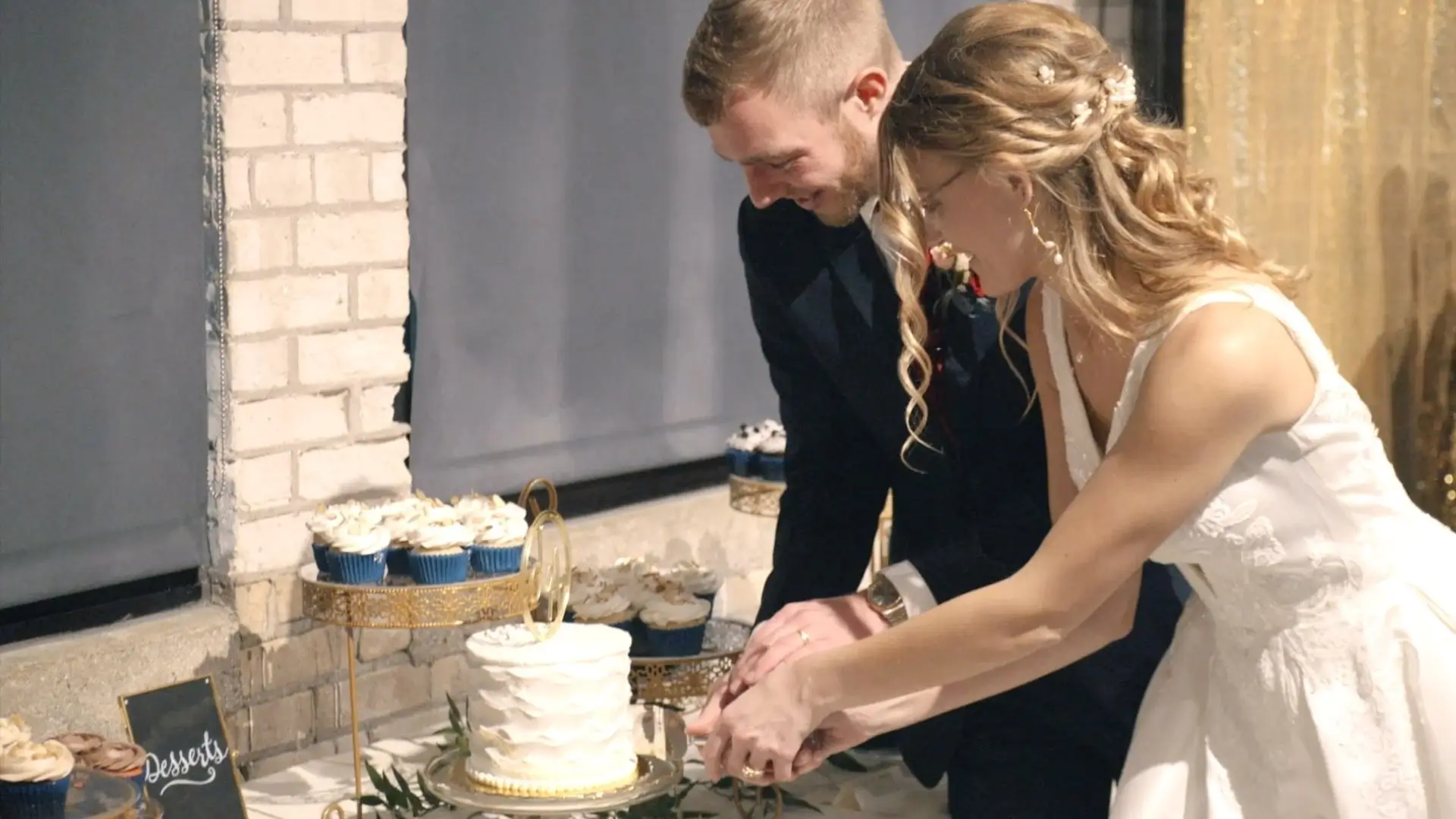Bride and groom, Julia and Andrew, cutting their wedding cake indoors. The dessert table has cupcakes with white icing and a small, elegantly decorated cake. The bride is in a white lace dress, and the groom wears a dark suit. 