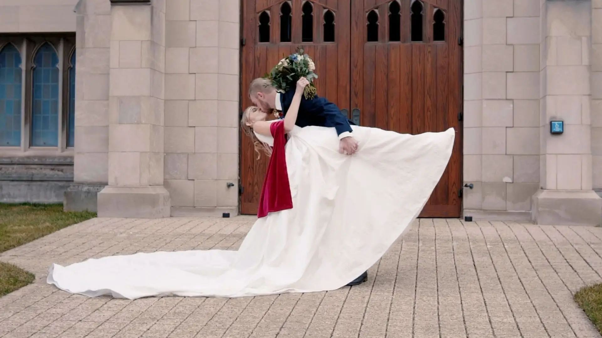 Andrew dips Julia while kissing outside a large church with wooden doors and stone walls in Holland. Julia wears a flowing white wedding dress with a long train, and Andrew is in a blue suit. Julia holds a bouquet of flowers.