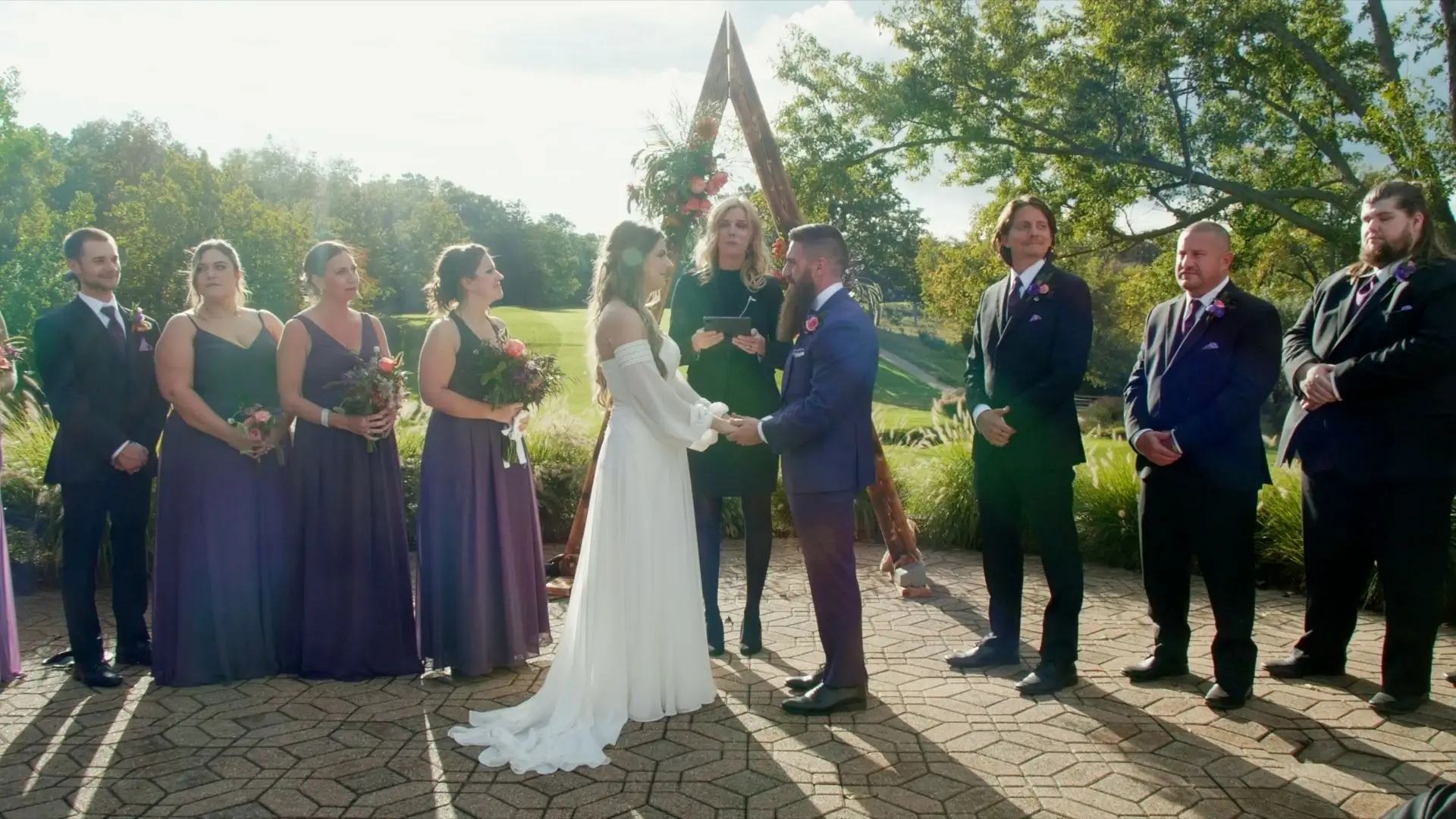 Emily and Tony exchanging vows outdoors at Plum Hollow, with their bridesmaids and groomsmen in the background. Emily wears a white gown, and Tony is in a blue suit. A minister officiates the ceremony, surrounded by greenery and a triangular arch.