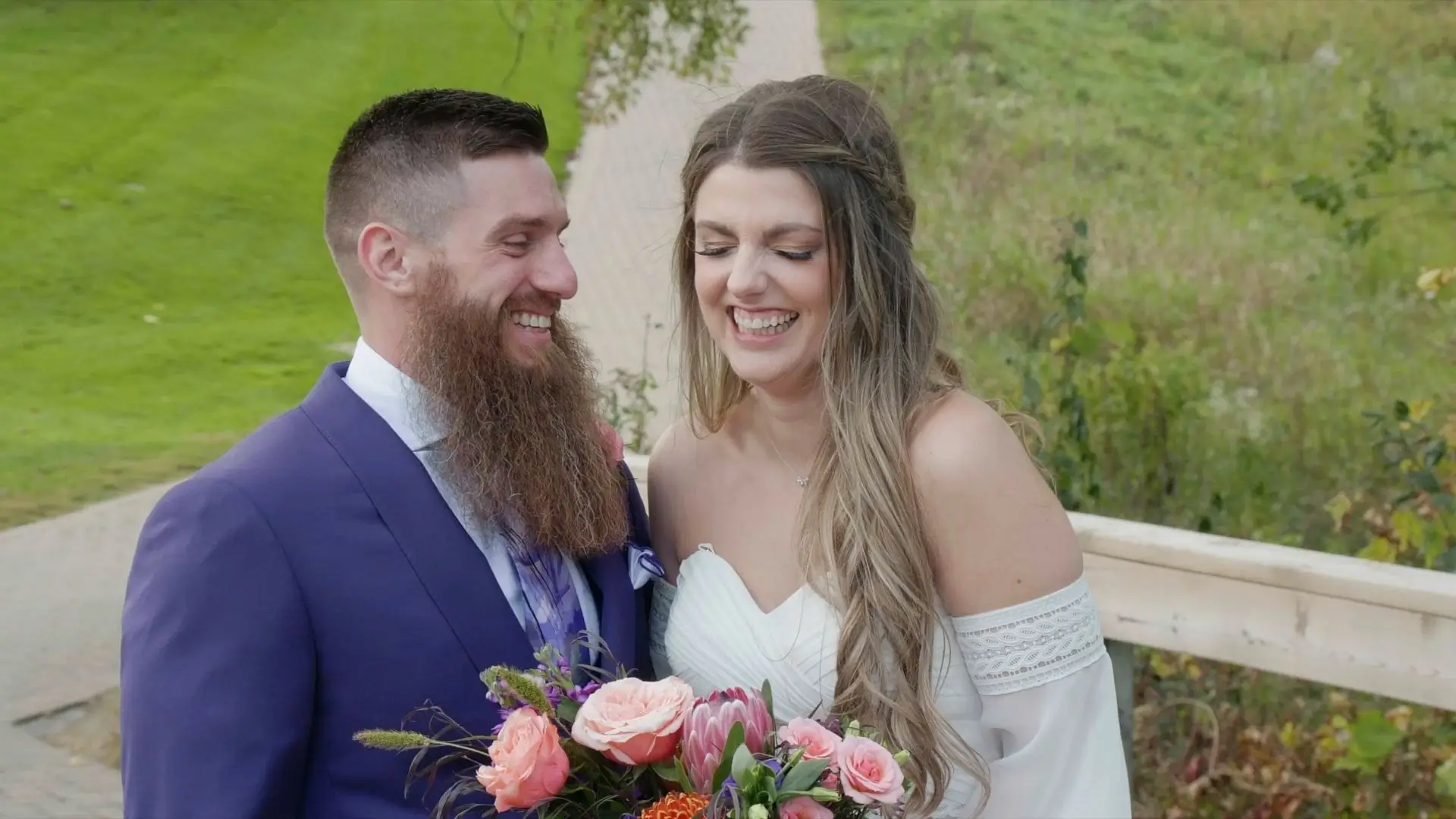 Emily and Tony, the bride and groom, are smiling at each other during their wedding in Southfield. Emily wears an off-shoulder white dress and holds a bouquet of pink and orange flowers. Tony is dressed in a purple suit.