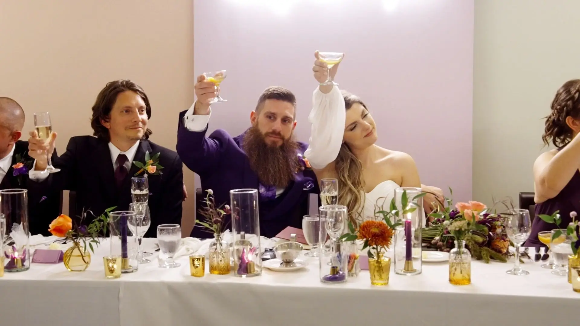 In this lively still from Emily and Tony's wedding video, the couple and their friends raise glasses in a celebratory toast at Plum Hollow Country Club in Michigan, surrounded by vibrant floral arrangements and joyful laughter.