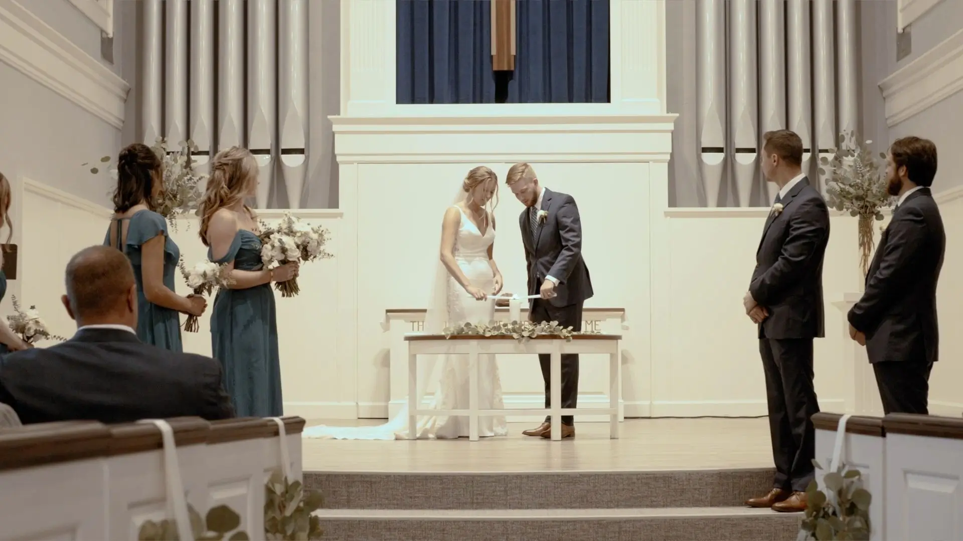 Bride Sophie and groom Jake perform a unity ceremony at their wedding in Pillar Church. Bridesmaids in teal dresses and groomsmen in black suits stand beside them, holding bouquets. Guests watch from the pews.