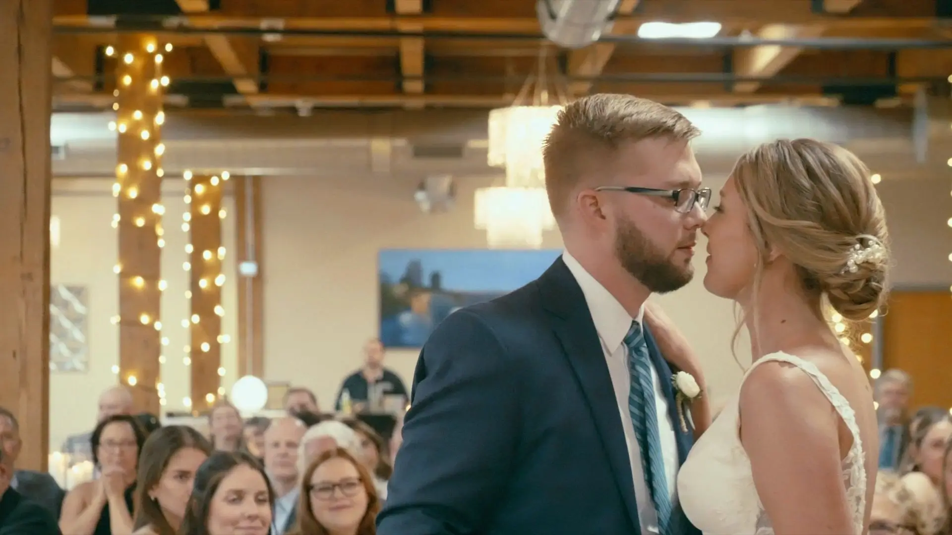 Bride Sophie and groom Jake share a tender moment at their wedding, standing close with foreheads touching inside Pillar Church decorated with strands of lights as guests look on.