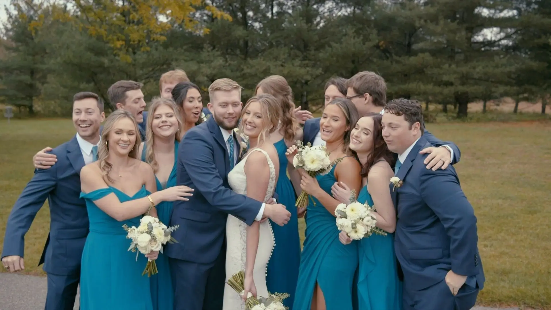 A joyful moment captured by the wedding videographer shows bride Sophie and groom Jake surrounded by their wedding party. Bridesmaids in teal dresses and groomsmen in blue suits smile and laugh together outdoors with greenery in the background.