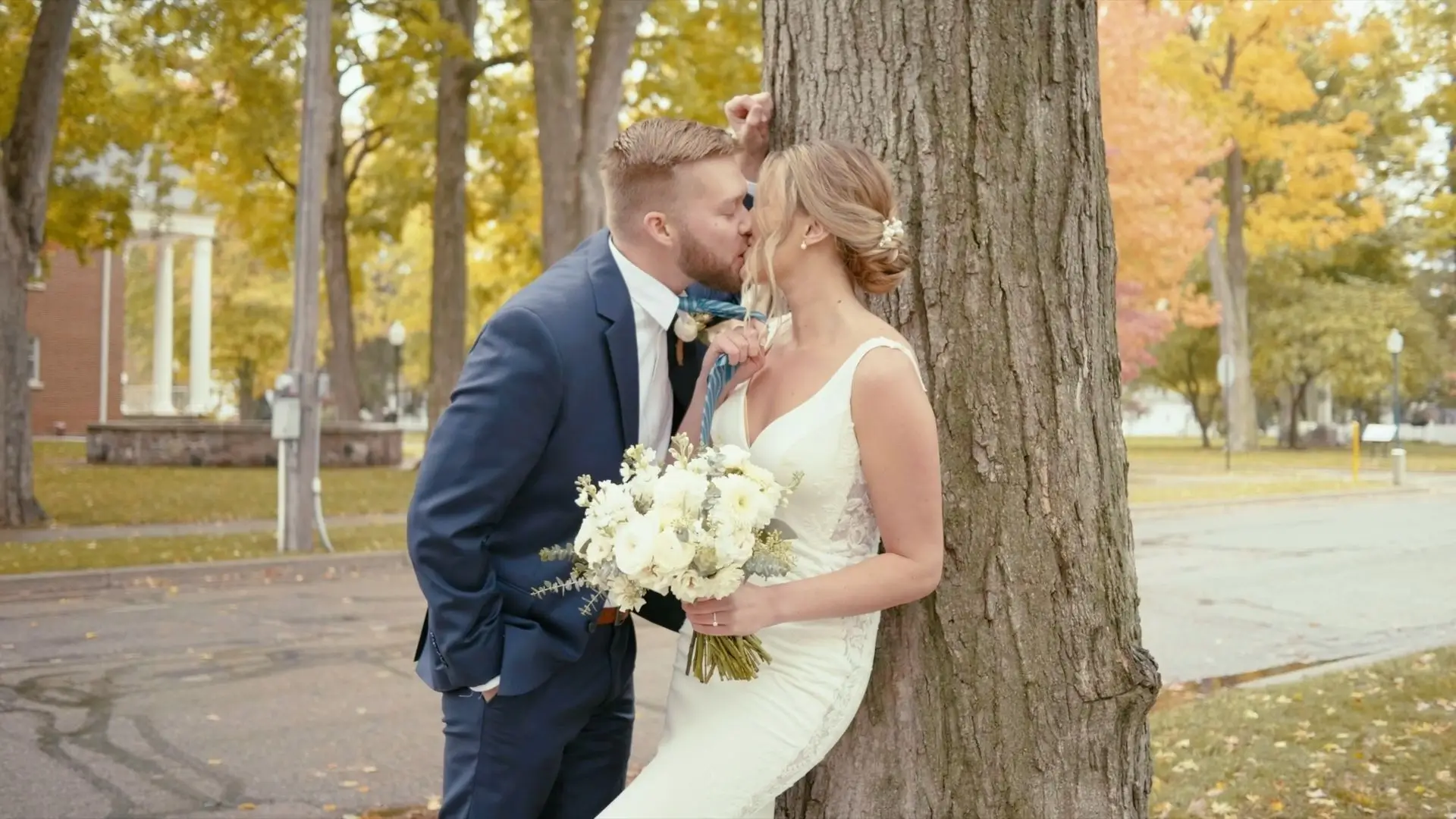 Jake and Sophie share a kiss in front of a large tree on their wedding day. Sophie, in a white gown and holding a bouquet, leans against the tree while Jake, in a navy suit, leans in. The background features fall foliage and a Pillar Church.