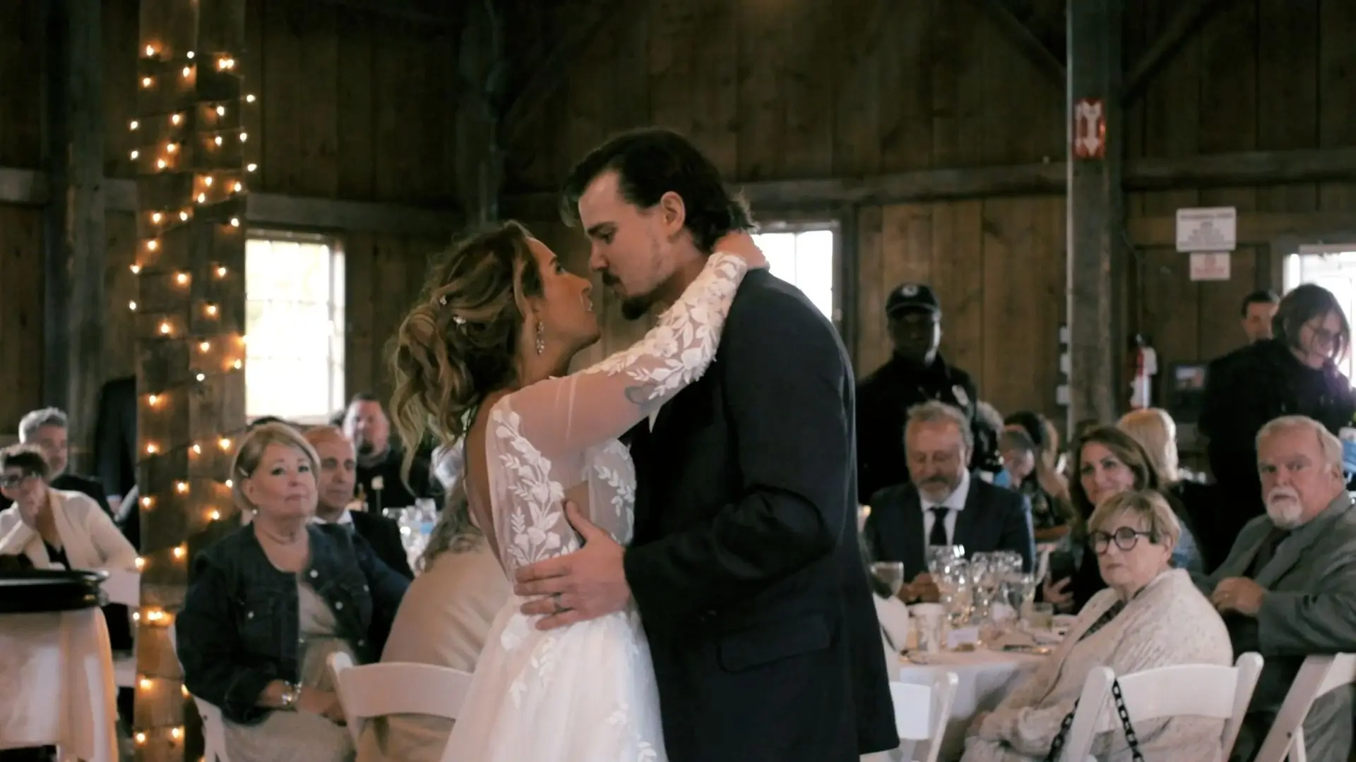Alyssa and Logan share their first dance as newlyweds at the Garvey Family Wedding Barn, with family and friends watching. The rustic venue is decorated with string lights, creating a warm, intimate atmosphere.