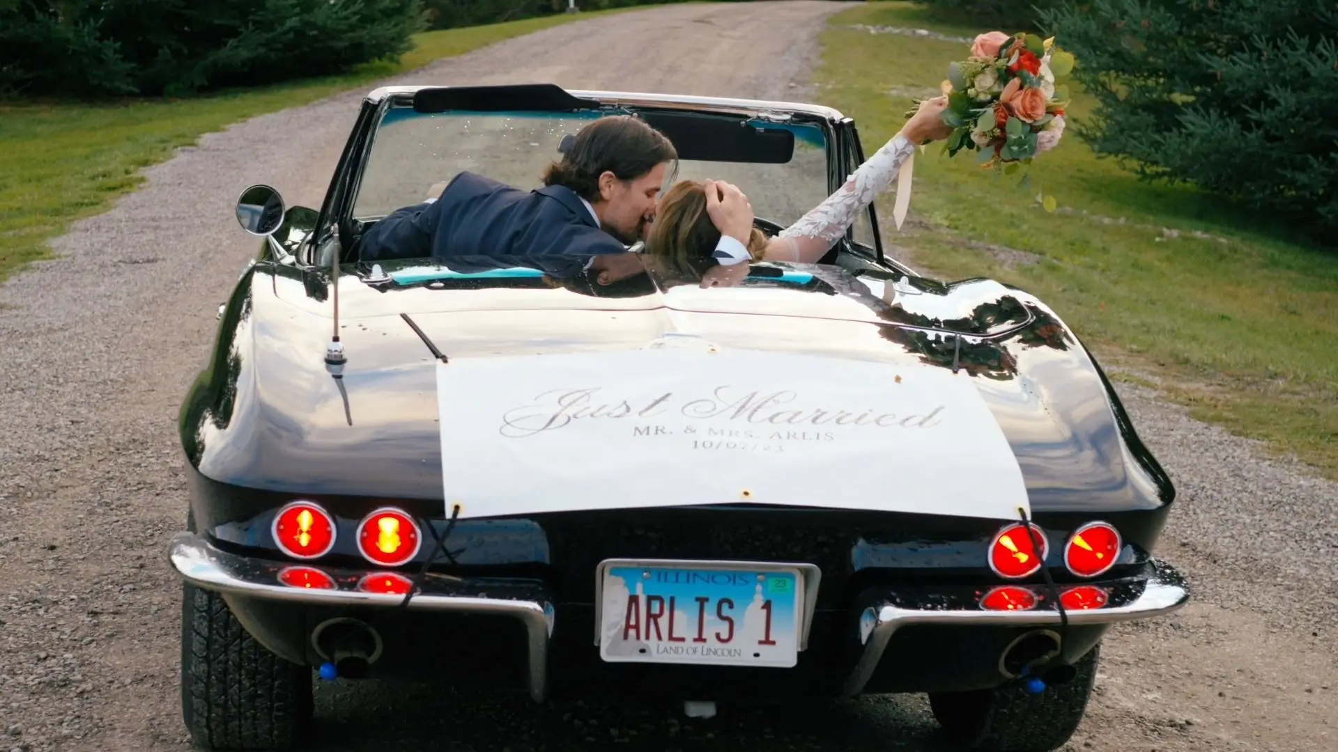 Newlyweds Alyssa and Logan are kissing in the front seat of a classic black convertible with a "Just Married" sign. Alyssa is holding a bouquet in the air. The car is on a gravel road surrounded by greenery at a wedding venue in Northern Michigan.