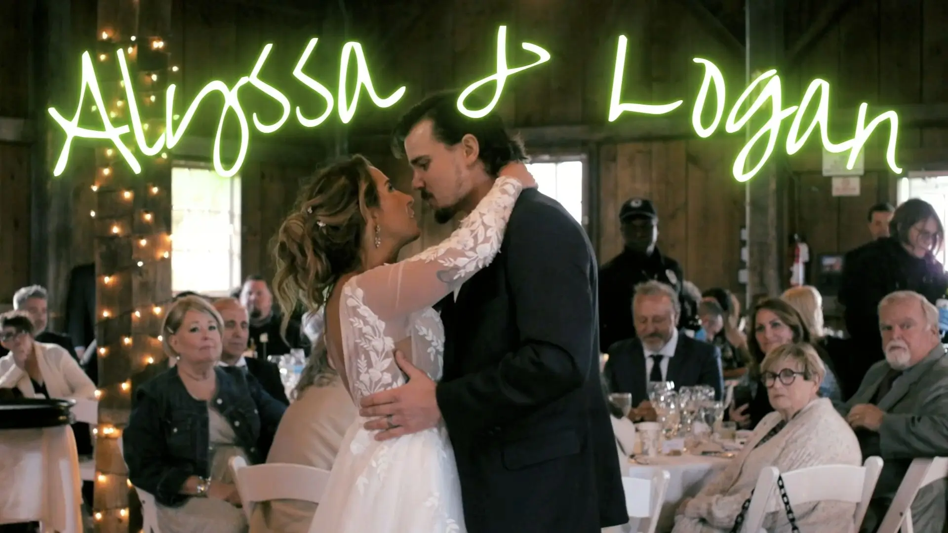Alyssa and Logan share a romantic first dance at Garvey Family Wedding Barn in Williamsburg. String lights wrap around wooden beams, and guests at tables watch and enjoy the moment. The words "Alyssa & Logan" are written in neon green above the couple.