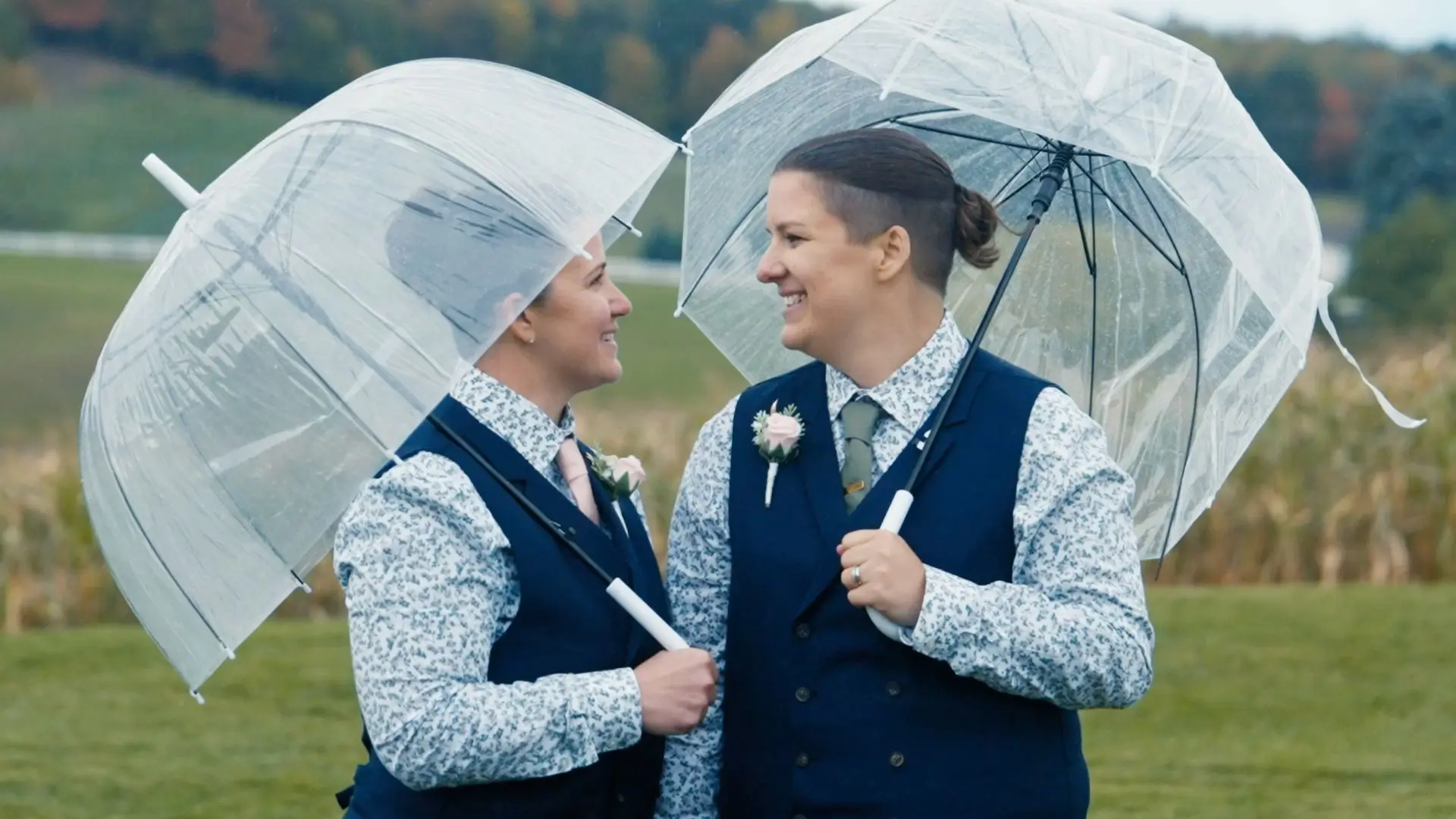 In this wedding highlight, Alexis and Carly smile at each other with clear umbrellas on a rainy day in Northern Michigan, wearing matching floral shirts and navy vests, captured candidly by their videographer.