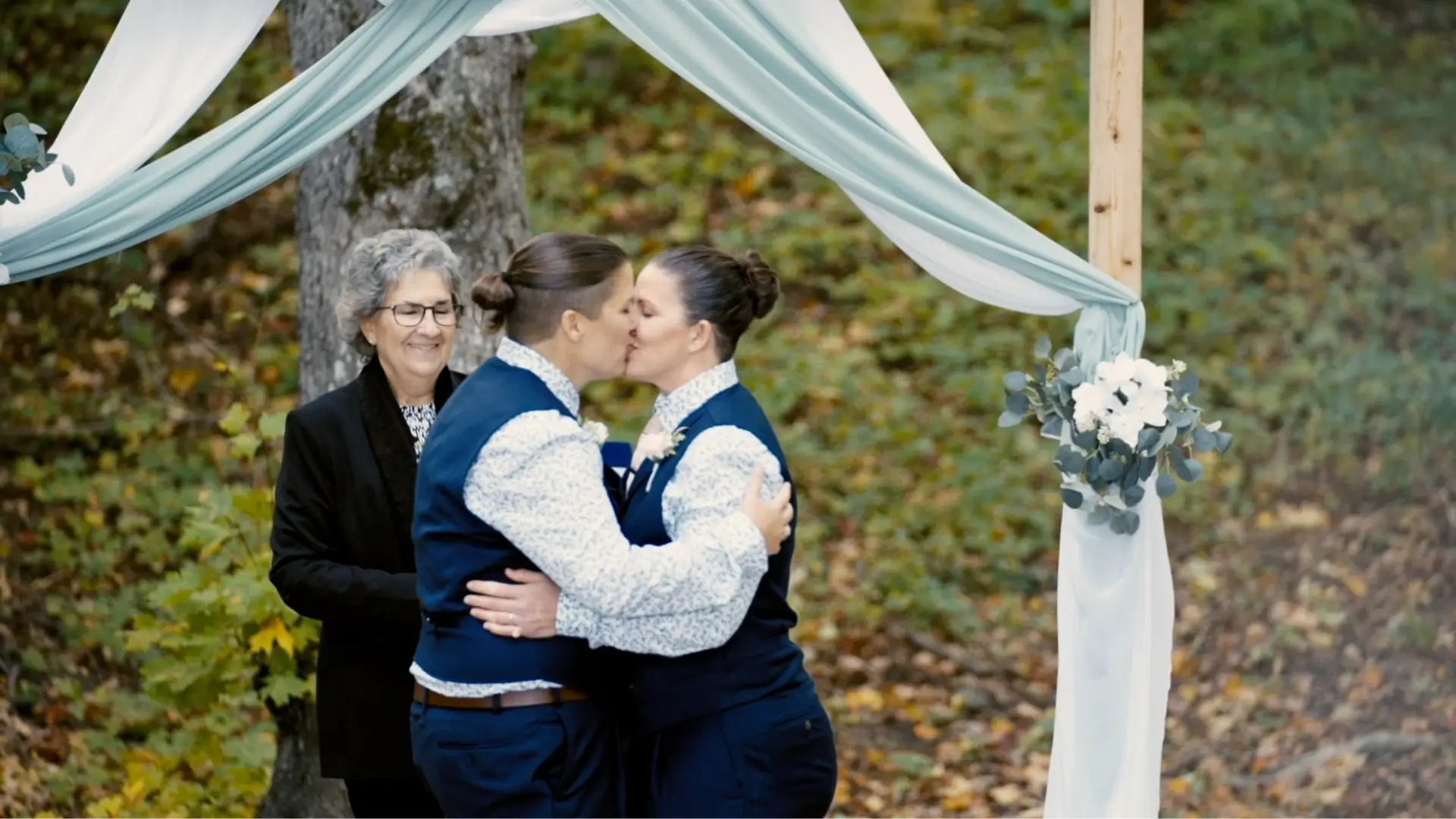 A couple, Alexis and Carly, share a kiss under an arch with green and white drapery in a lush Michigan forest. Both are wearing blue vests and patterned shirts. An officiant stands nearby smiling.