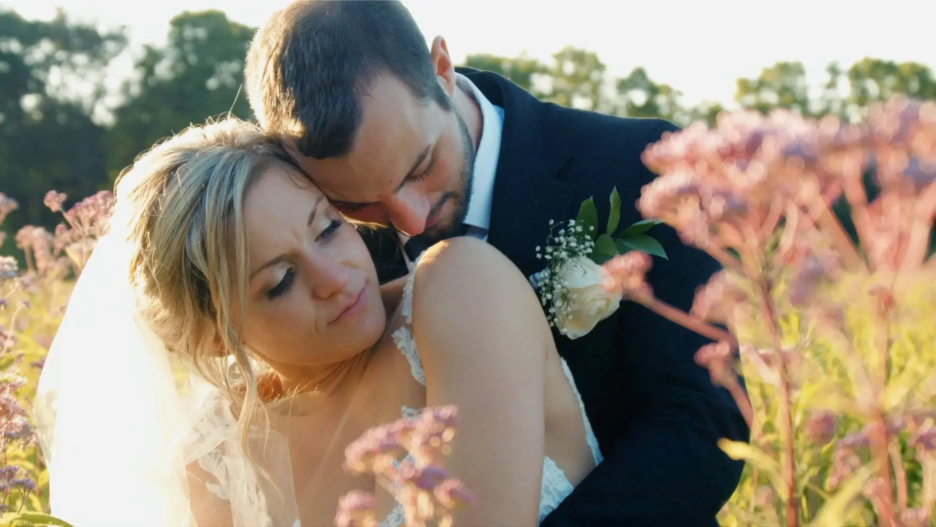 Bride Casey and groom Andrew embrace in a field of flowers at sunset, captured by their wedding videographer. Casey wears a veil and lace dress, while Andrew is in a suit with a boutonnière. Flowers and trees fill the background.