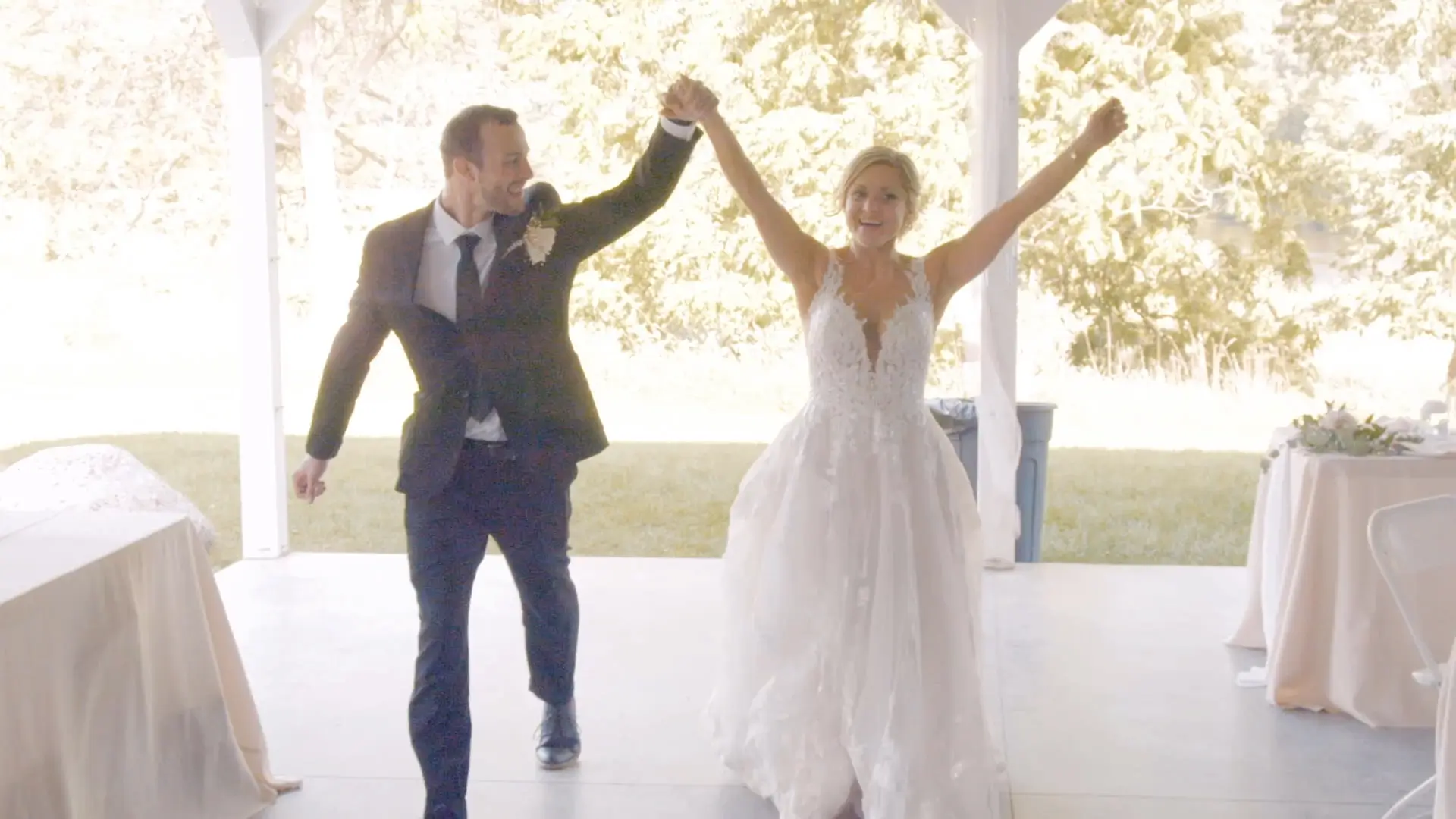 Casey and Andrew hold hands and cheer in celebration during their wedding in Lowell. The bride looks radiant in her lace gown while the groom beams in his dark suit. Tables and green trees are visible in the background.