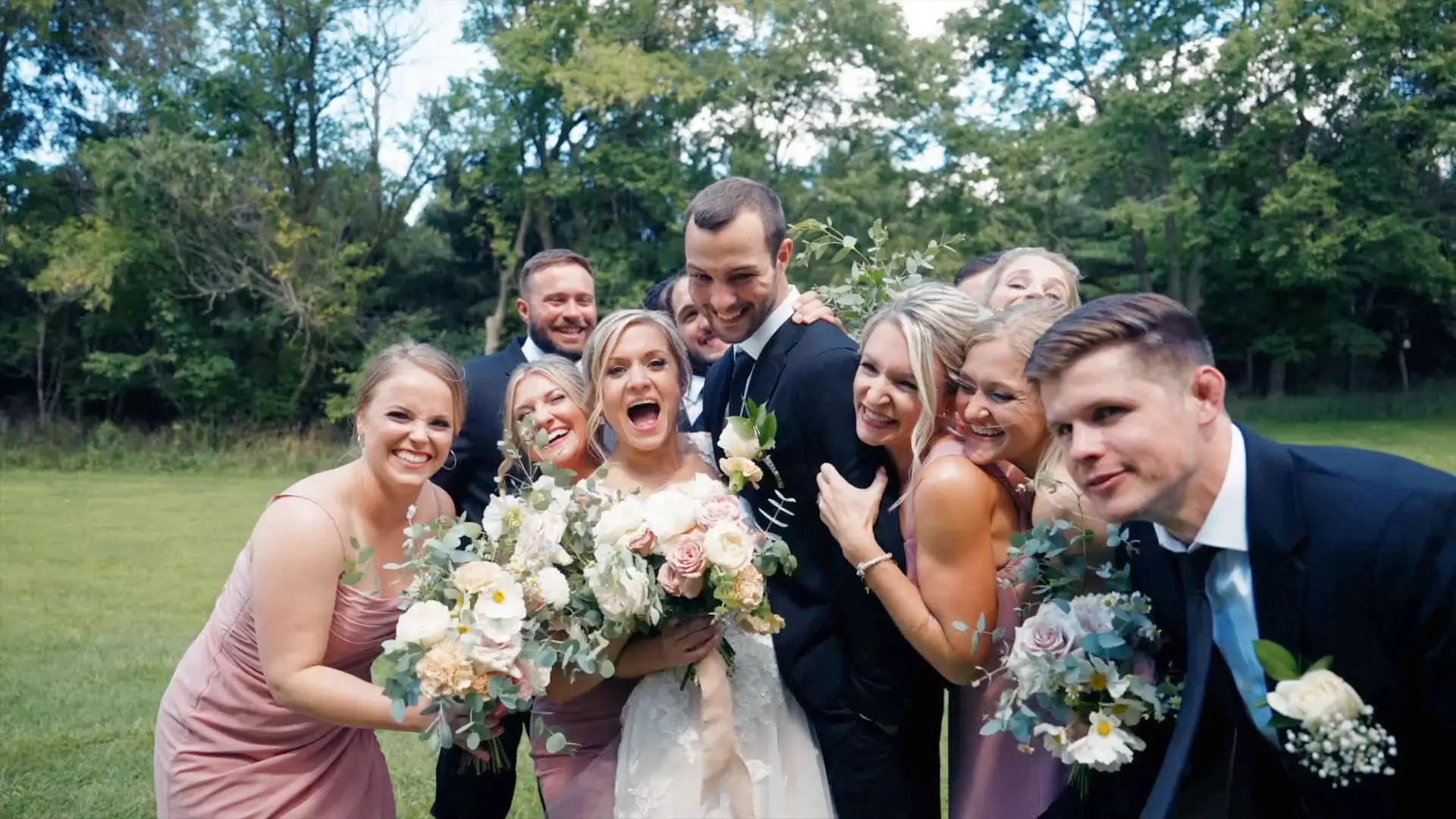 The bride, groom, and wedding party smile and laugh in a group photo outdoors. The bride holds a bouquet of flowers as they pose for their wedding videographer amid a lush, green backdrop in Lowell.