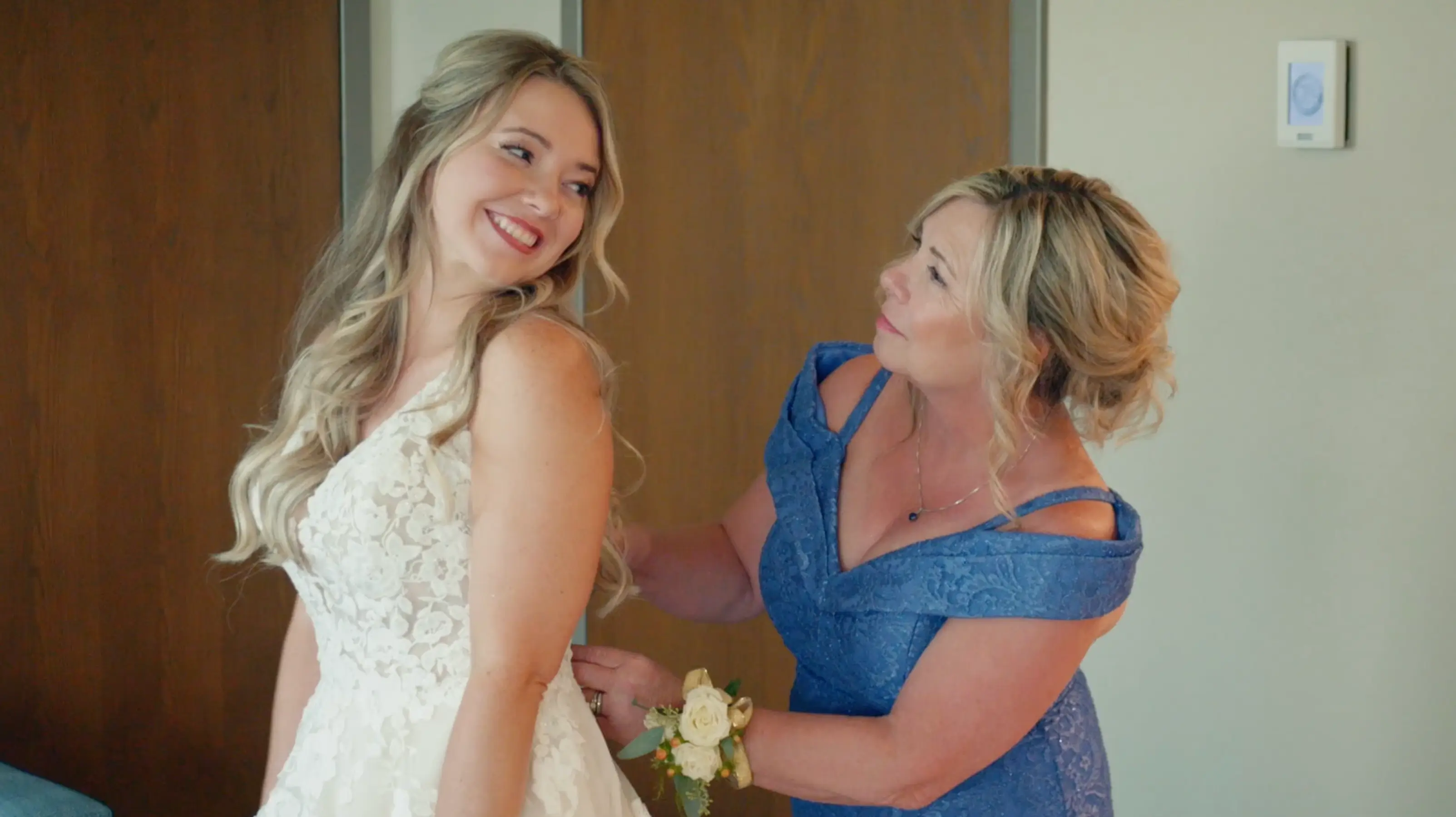 Nicole in her wedding dress smiles joyfully as her mother, dressed in a blue lace gown, helps button her dress at the Grand Traverse Resort & Spa in Traverse City, Michigan.