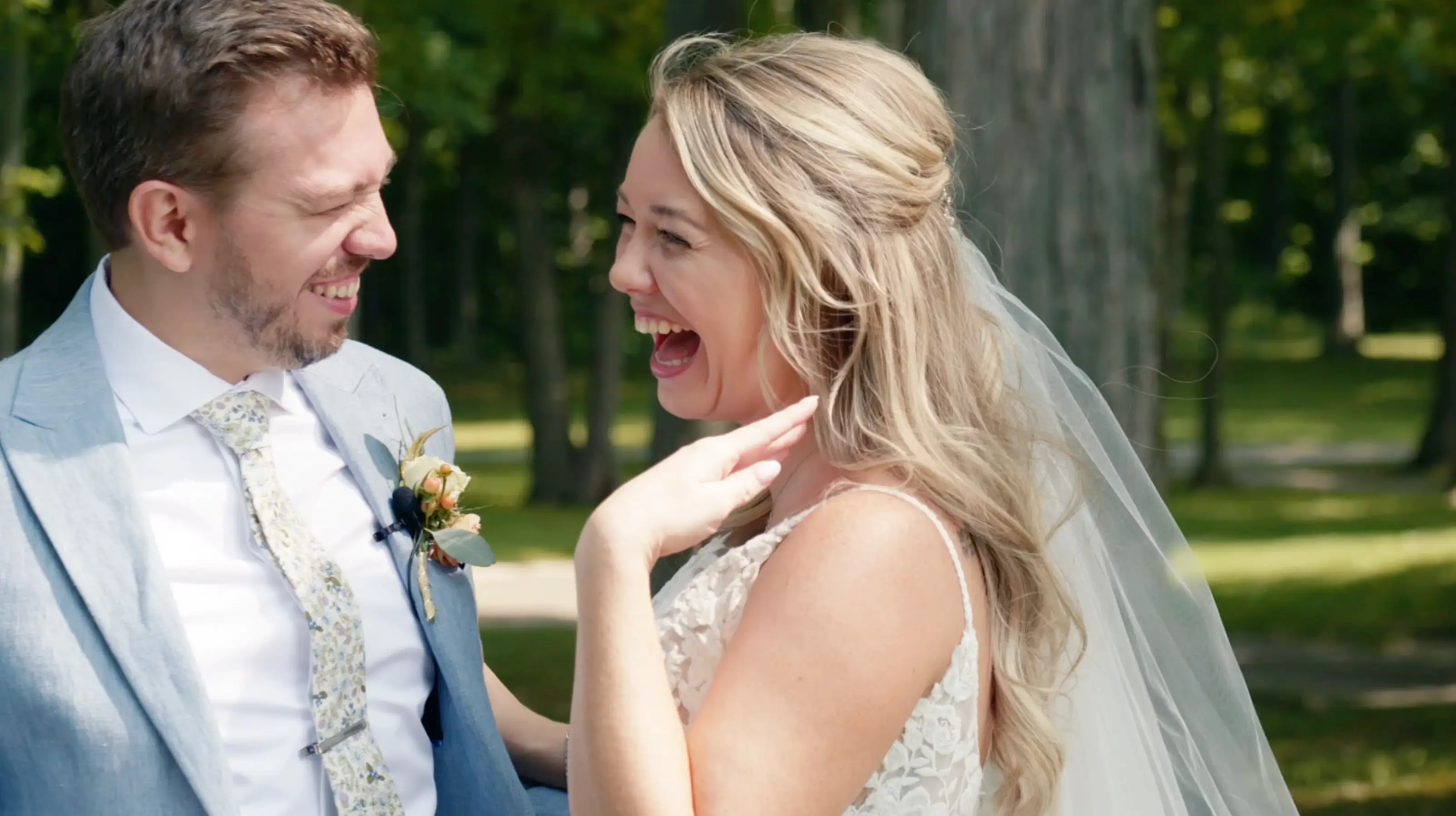 A joyful bride and groom sharing a candid moment of laughter outdoors. The bride, wearing a lace dress and veil, has her hand raised playfully. The groom, dressed in a light suit, looks at her affectionately.