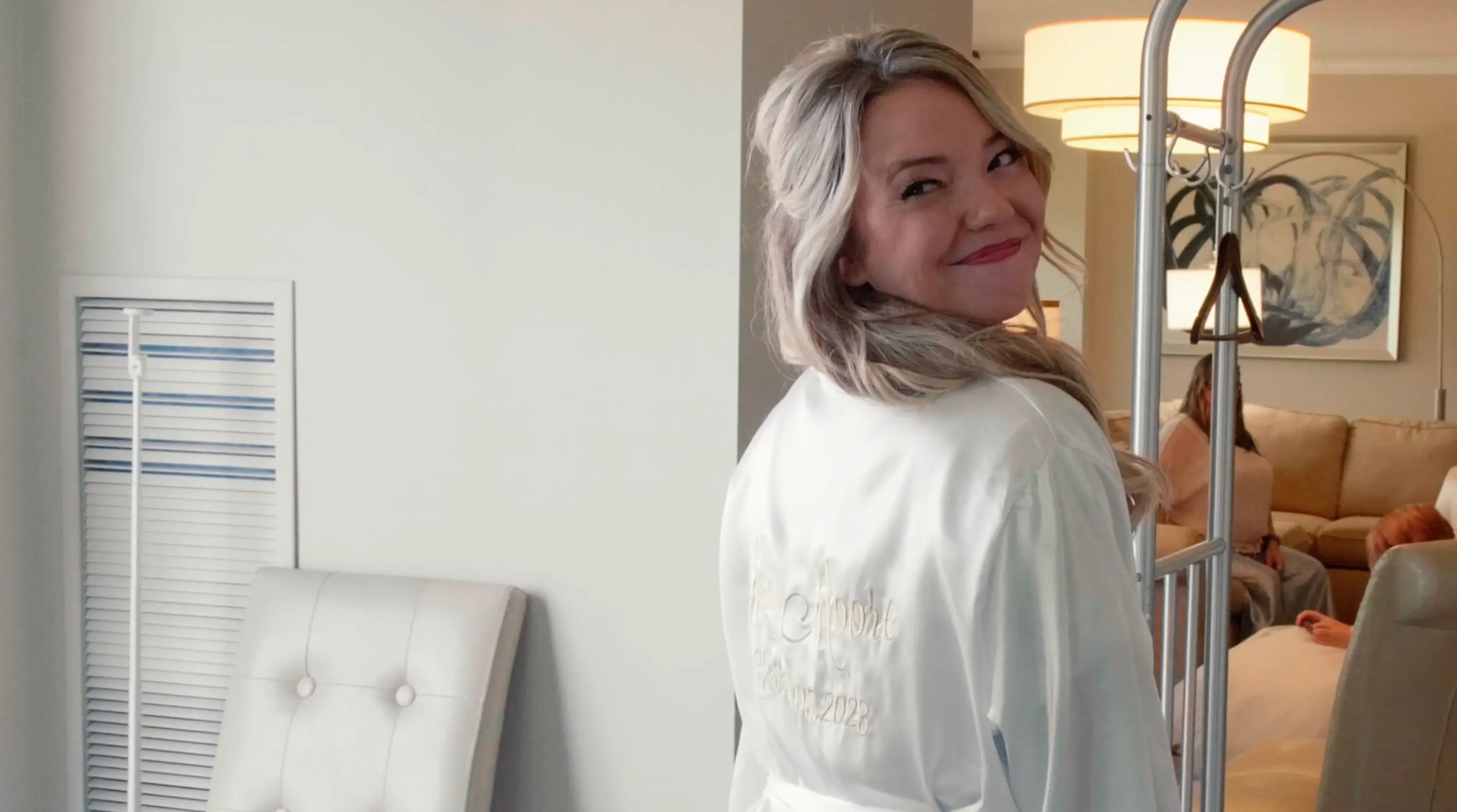 Nicole getting ready for her wedding at Grand Traverse Resort & Spa in Traverse City, Michigan, smiling as she turns back. She wears a white robe with embroidery, and people are visible in the background in a cozy indoor setting.