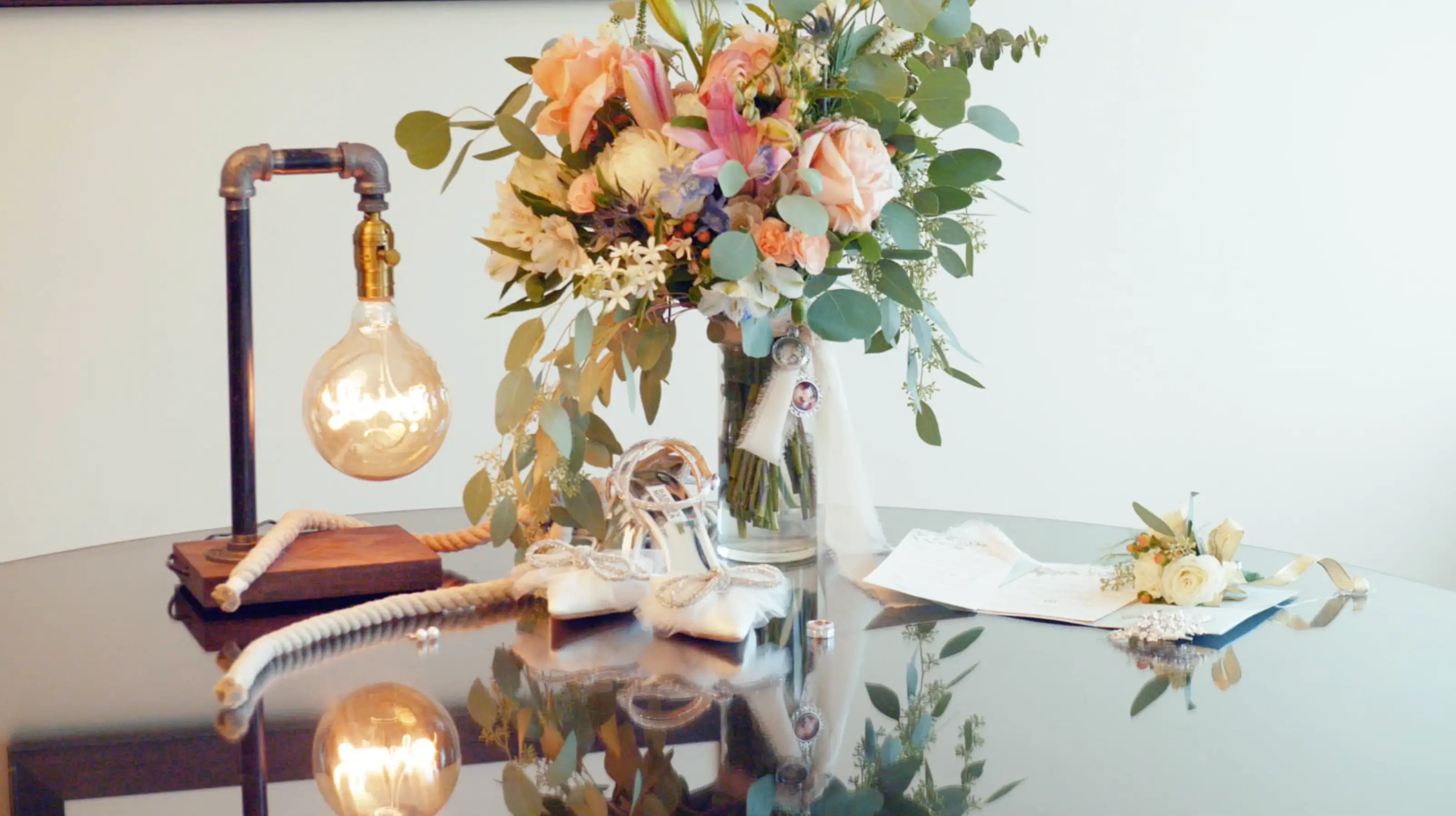 A round table holds a decorative light fixture, a bouquet of flowers, two photo charms, a pair of bridal shoes, a ring, and a white floral corsage on a folded paper.