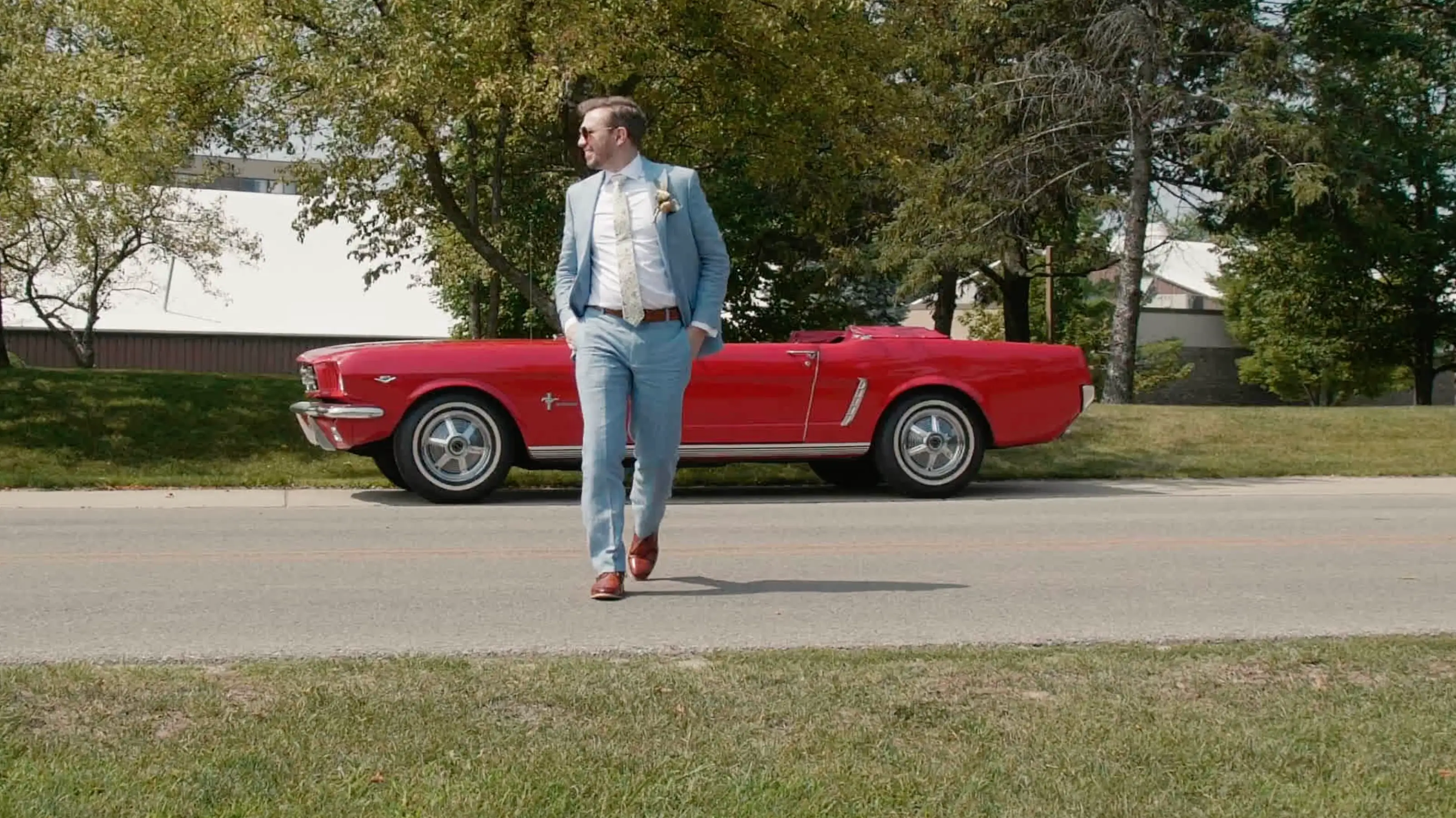James, the groom, wearing a light blue suit with a boutonnière walks confidently across a road in front of a classic red convertible, surrounded by trees in Traverse City.