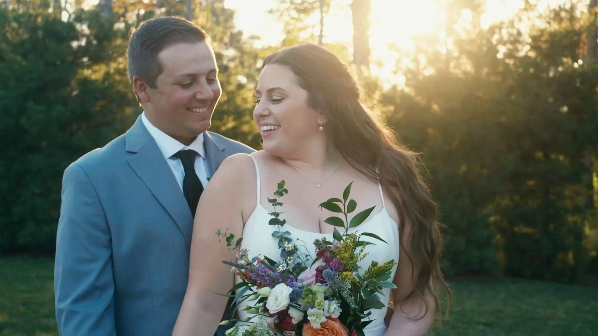 Bride Ryleigh and Groom Nick share a candid moment at their outdoor wedding at Black River Barn. They smile at each other, with Ryleigh holding a colorful bouquet as the sun sets behind them, creating a warm, golden glow.