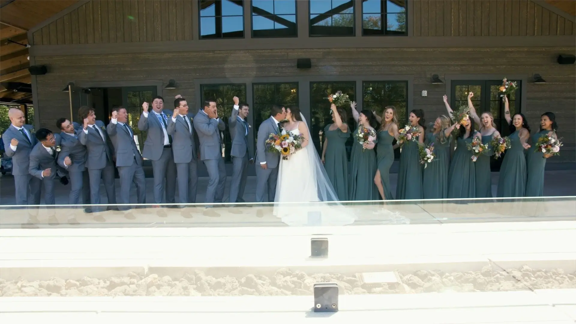 A wedding party stands in front of Black River Barn in South Haven, Michigan. The bride, Ryleigh, and the groom, Nick, share a kiss in the center while bridesmaids in green dresses cheer and groomsmen in grey suits celebrate joyfully on either side.