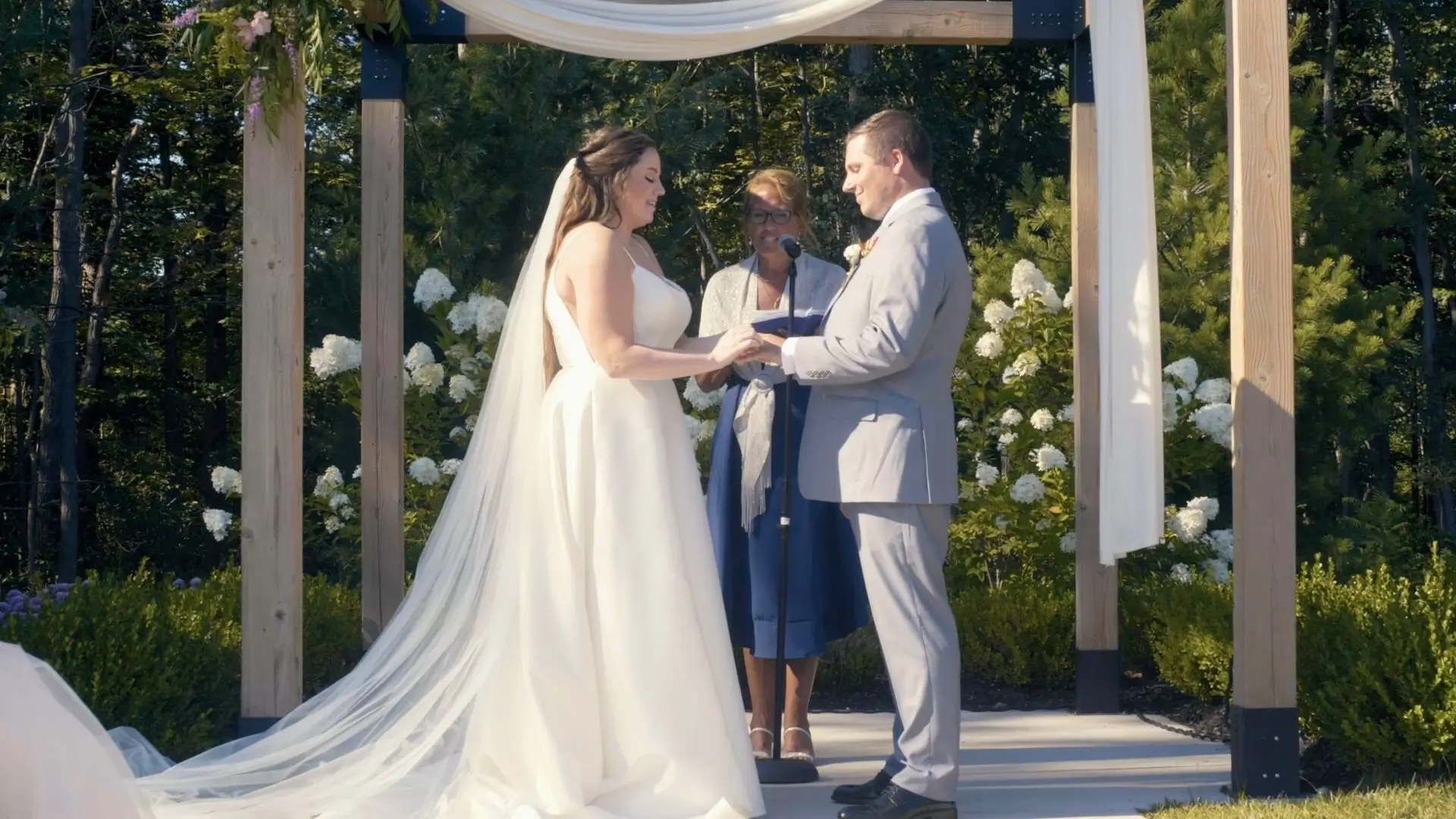 Bride and groom exchanging vows under a wooden arch draped with white fabric and decorated with flowers. The bride wears a flowing white dress with a long veil, and the groom is in a light grey suit. An officiant stands behind them.