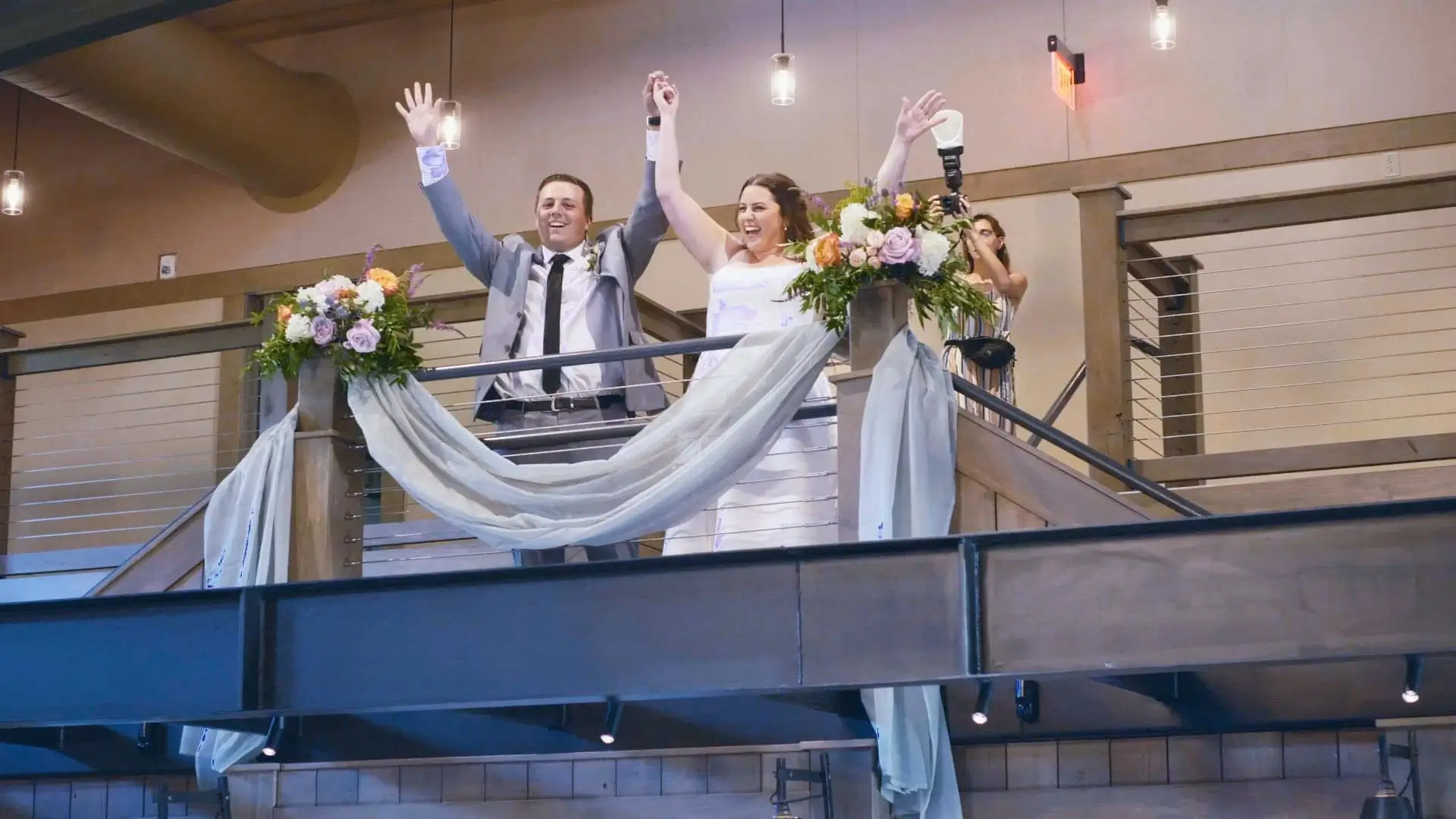 Bride Ryleigh and groom Nick celebrate by raising their hands on a decorated balcony at Black River Barn in South Haven, Michigan. A photographer captures the moment from the side, while floral arrangements and draped fabric adorn the railing.