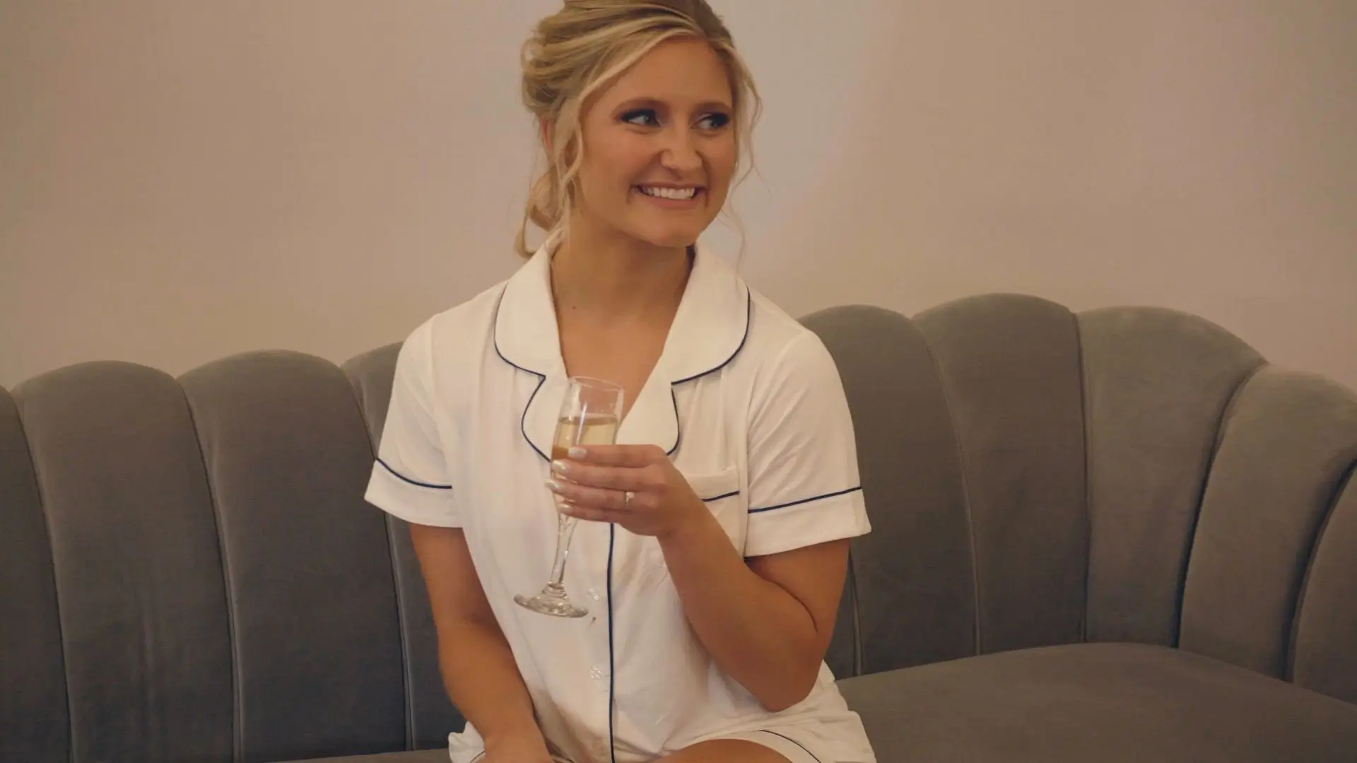 A joyful bride, Bethany, sits on a plush couch in Michigan, holding a champagne glass, captured in this candid moment by the wedding videographer.