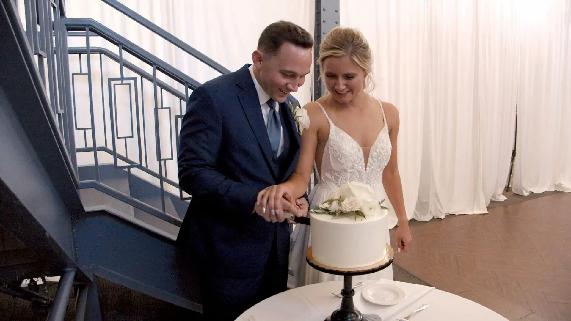 In a candid moment from Bethany and Nick's wedding video, the couple cuts their wedding cake in front of a staircase at Clearwater Place Events in Grand Rapids, Michigan. The bride wears a flowing white gown, and the groom wears a navy suit.